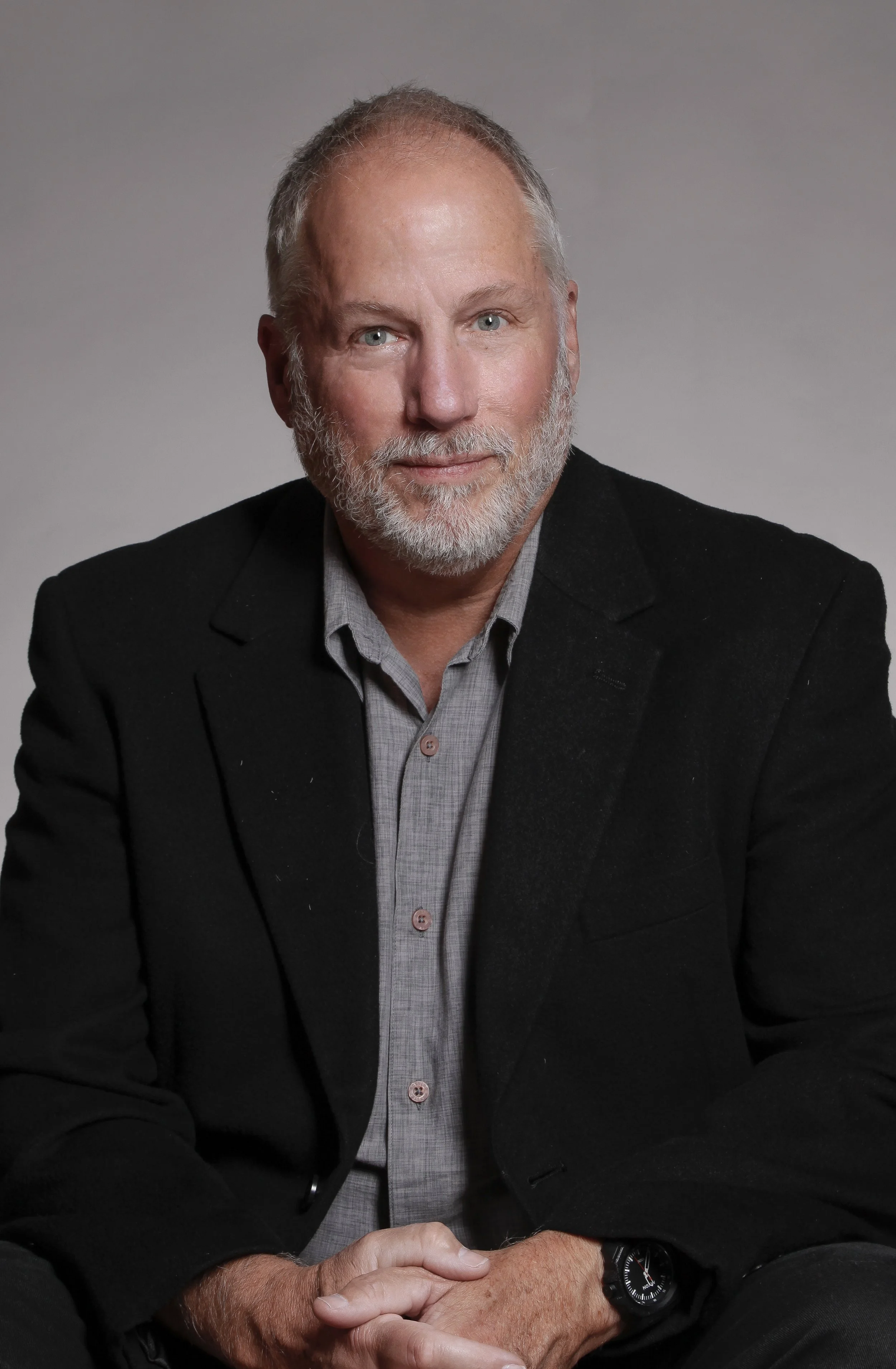 Headshot of an older man with gray hair and beard, dressed in a black blazer and gray shirt, sitting with hands clasped, wearing a black watch, against a plain background.