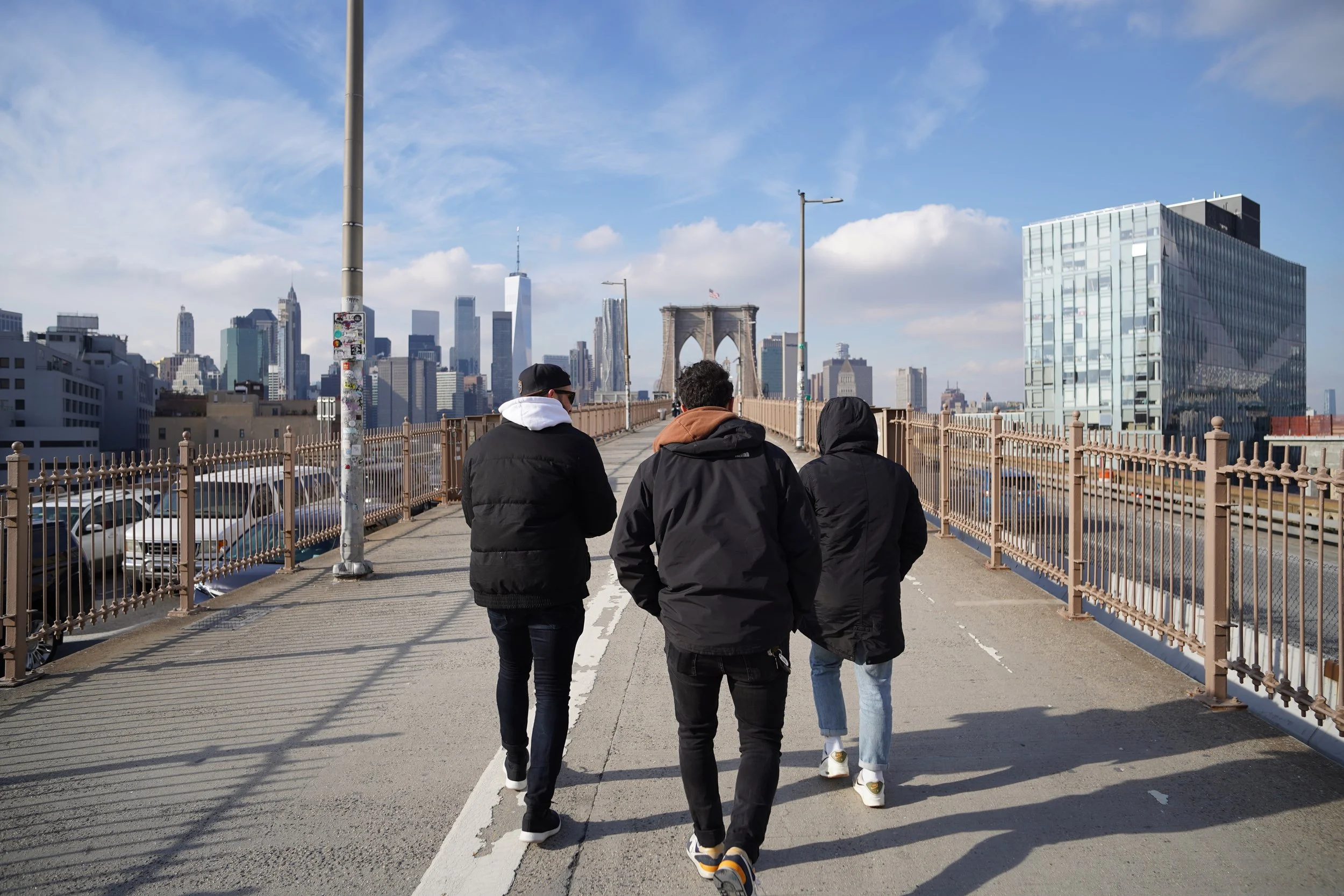 Three people walking on a bridge with a city skyline in the background, including the Brooklyn Bridge, tall skyscrapers, and a partly cloudy sky.