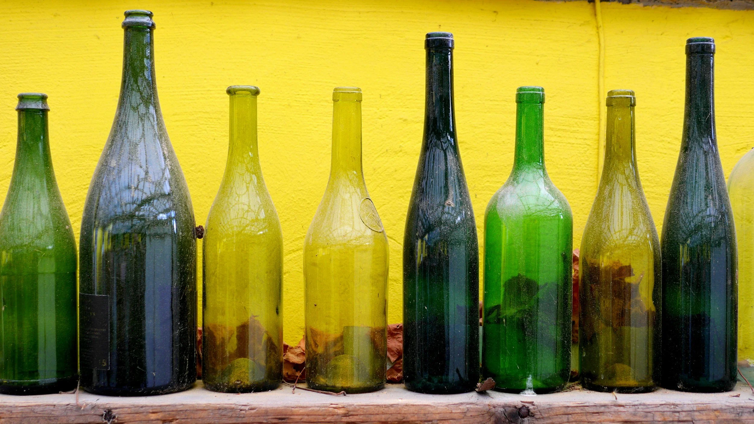 A row of empty glass bottles in various shades of green, yellow, and dark colors, lined up against a bright yellow textured wall.