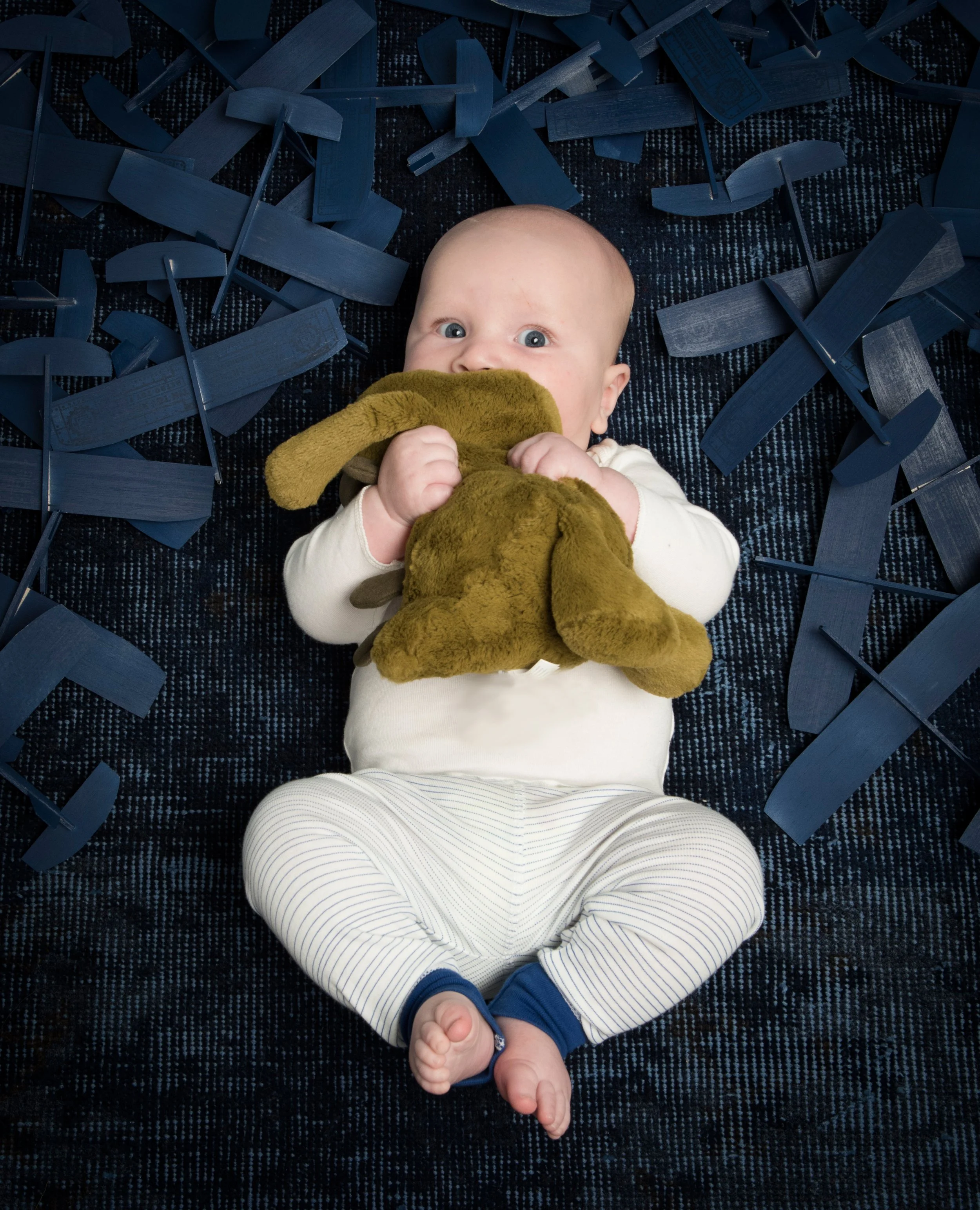 A baby lying on a dark textured carpet, holding a brown plush stuffed animal, wearing cobalt stripe pajama bottoms from the Little Auggie infant sleep set..