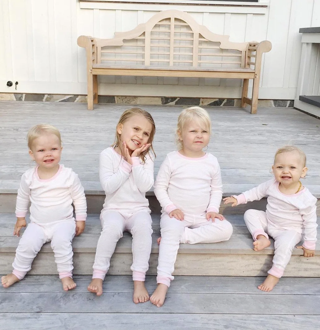 Four young children sitting on a wooden deck, dressed in matching pink and white pajamas by Little Auggie.