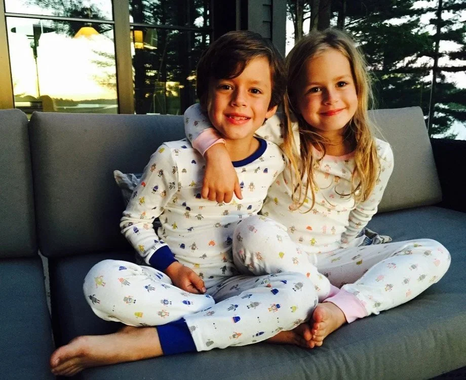 Two children, a boy and a girl, sitting on a gray couch in cotton pajamas.