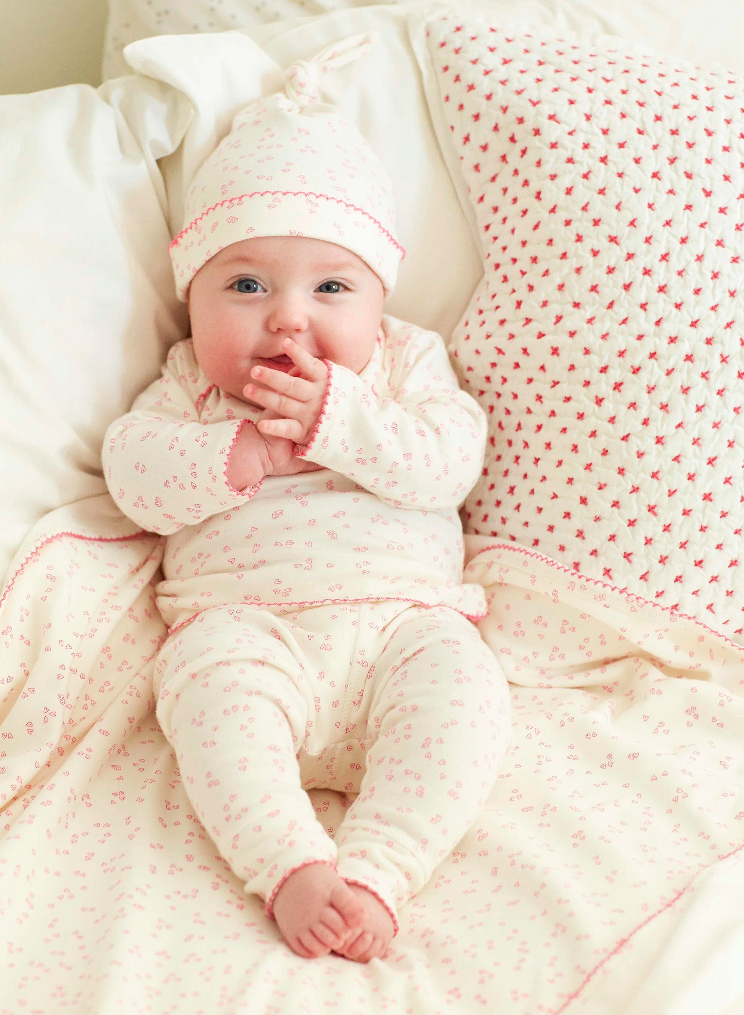 A baby lying on a bed, wearing pink and white pajamas with a matching hat and blanket from the organic cotton Little Auggie layette set.