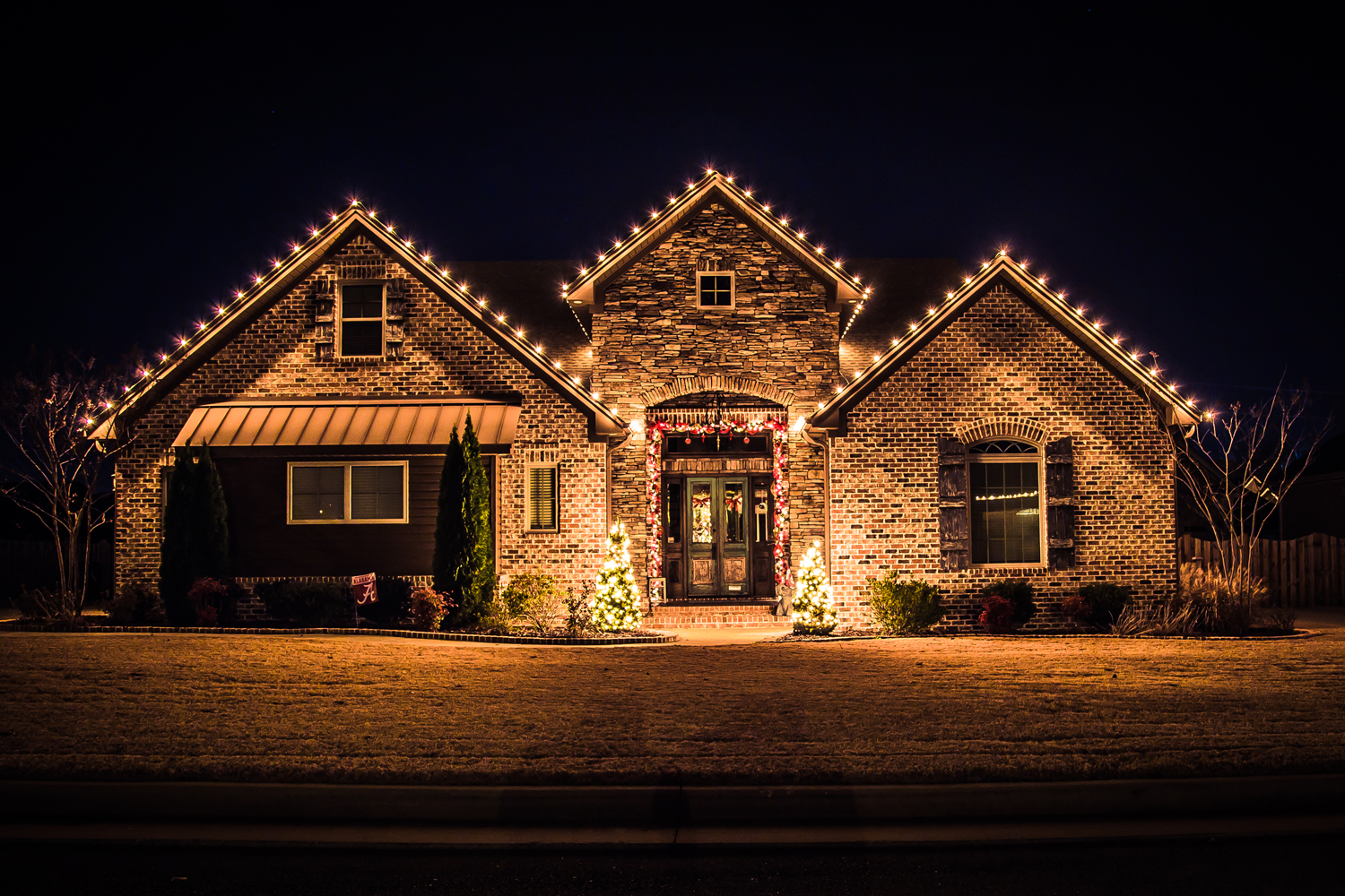 A brick house decorated with Christmas lights and ornaments at night, with trees and a lawn in front.