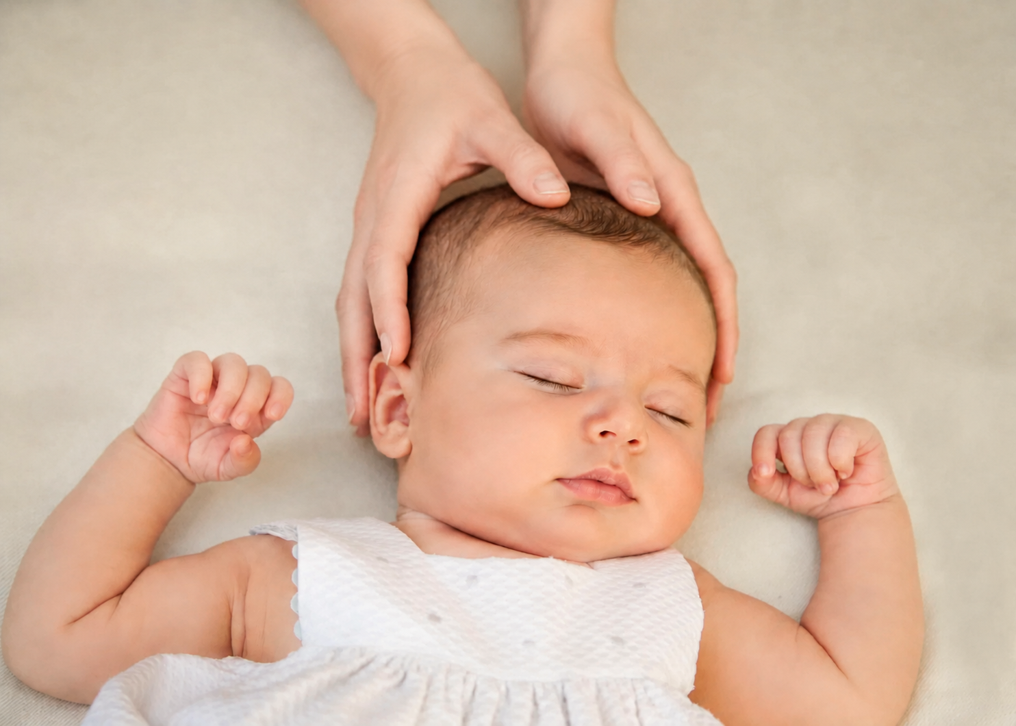 A baby with closed eyes, lying down in a white outfit, with hands loosely clenched, receiving a gentle head massage from adult hands on a soft surface.