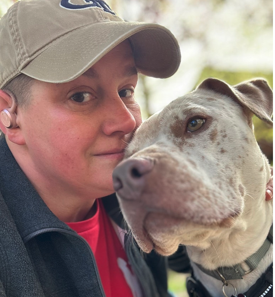A person and a large dog close together outdoors, with the person smiling slightly and the dog calmly looking at the camera.