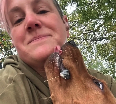A person with short hair wearing an olive green shirt, smiling as a dog licks their face in an outdoor setting with green trees. Dog walk