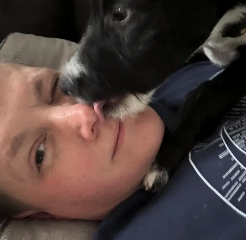 Person lying down with a black and white dog affectionately licking their face.