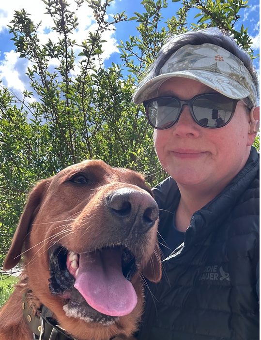 Person wearing sunglasses and a cap taking a selfie outdoors with a happy brown dog, trees, and a partly cloudy sky in the background.