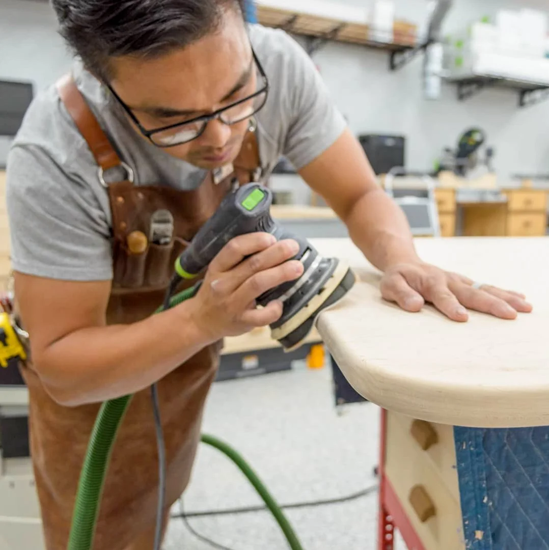 Huy wearing glasses and a leather apron is sanding a wooden piece with a power sander in a woodworking shop.