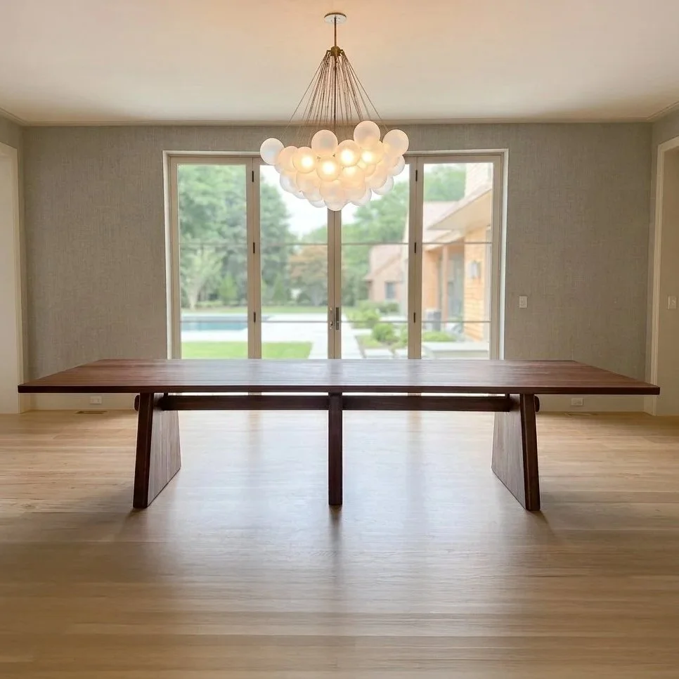 Handmade walnut dining table displayed in a bright modern dining room with large glass doors and a sculptural chandelier.