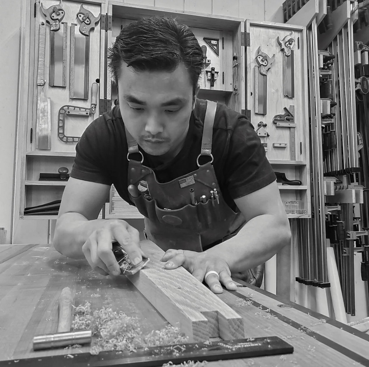 Huy Huynh using a hand tool to shape wood at a workbench in his workshop.
