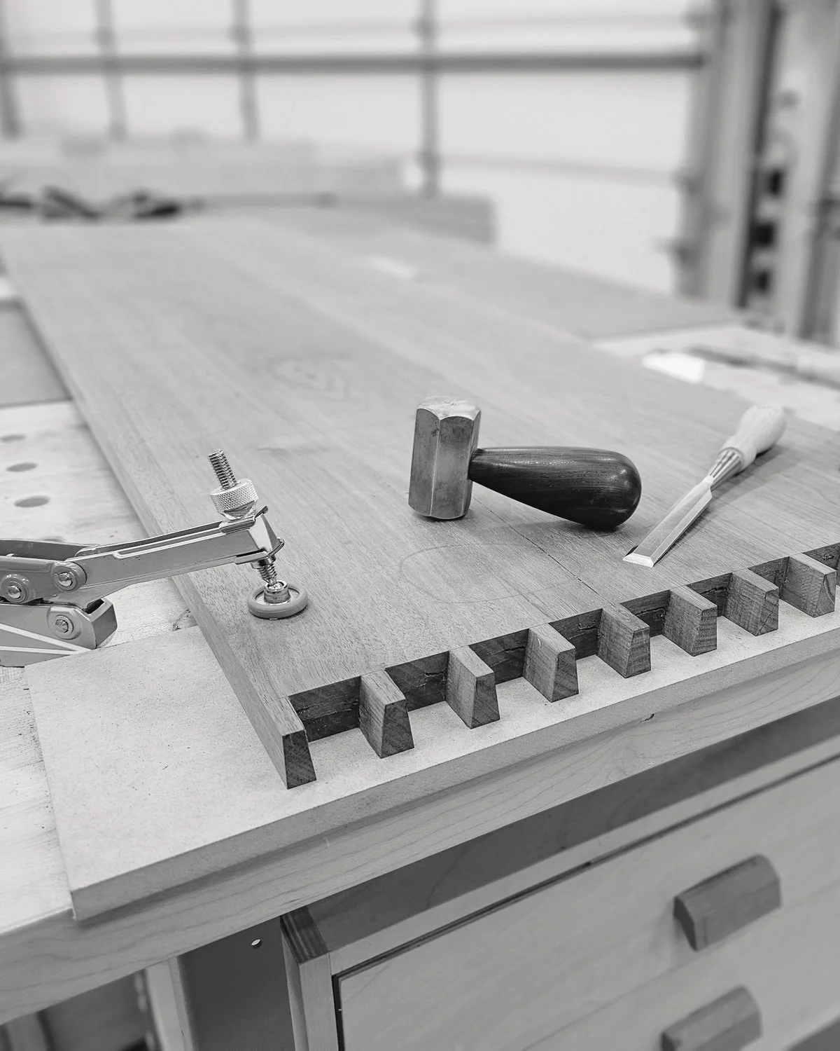 A woodworking workbench with tools including a hammer with a dark handle, a chisel, a screwdriver, and a clamp, along with a piece of wood with finger joints in a woodworking shop.