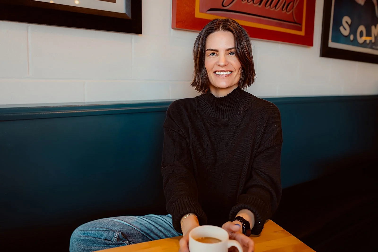 A smiling young woman with shoulder-length brown hair, wearing a black turtleneck sweater, sitting at a wooden table in a cafe, holding a white mug with a beverage, with framed artwork on the wall behind her.