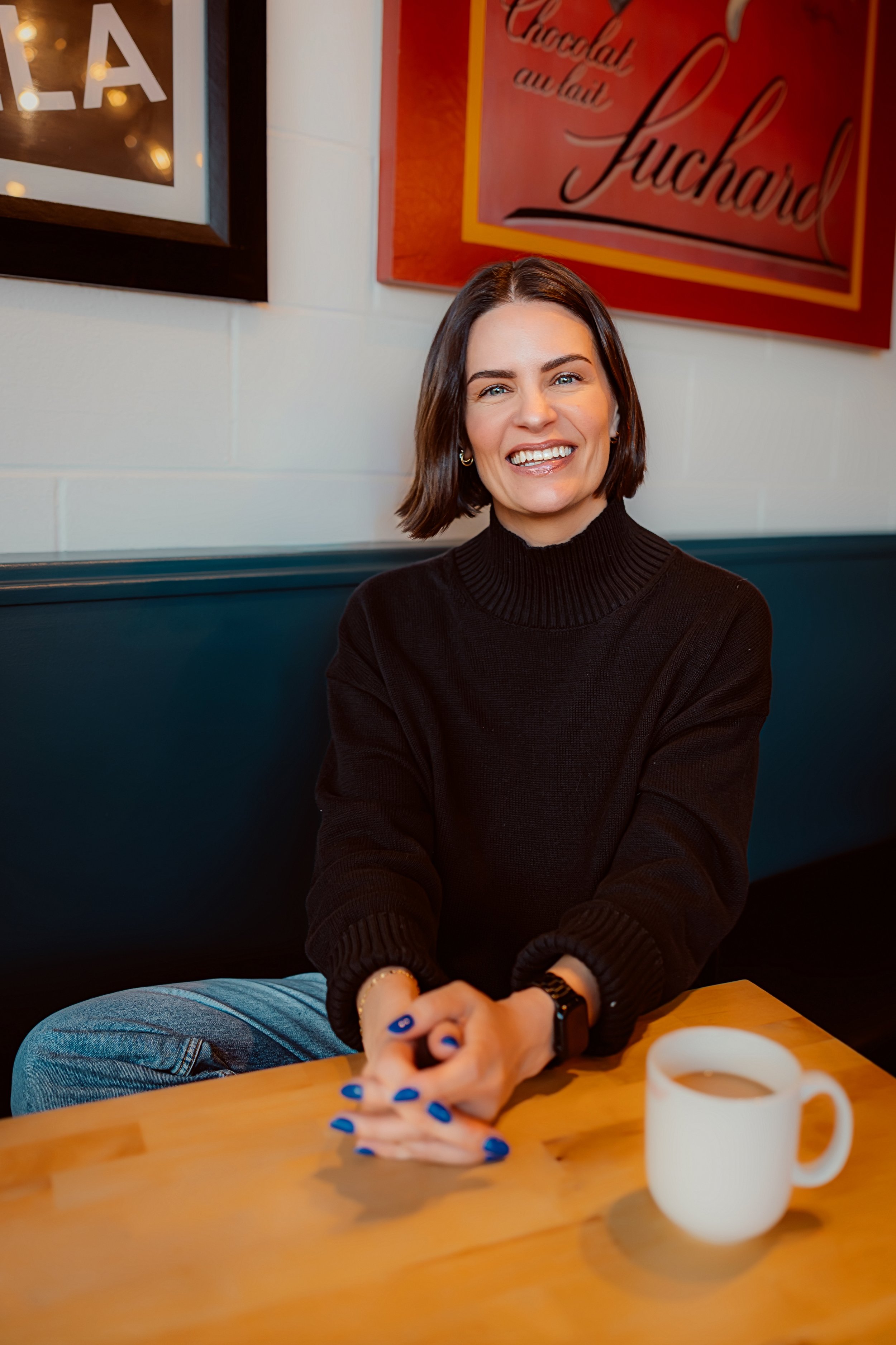 A woman with shoulder-length brown hair, wearing a black turtleneck sweater, smiling in a restaurant with a blue booth and white brick wall with colorful signs in the background. A white mug is on the wooden table in front of her.
