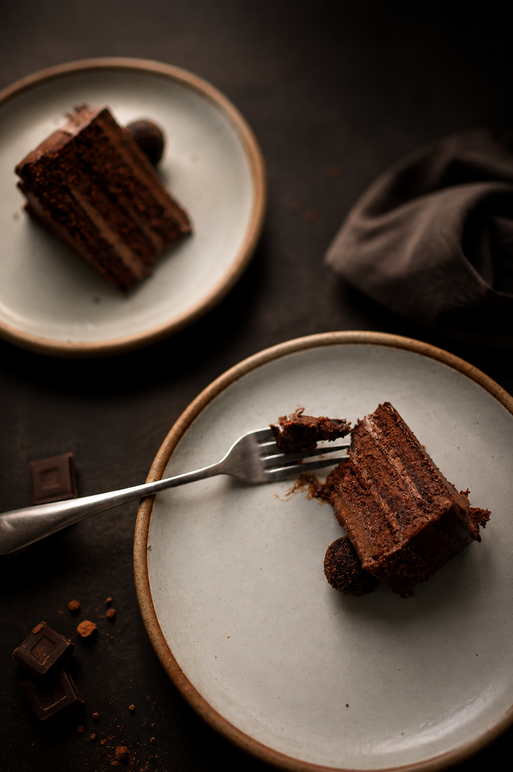 Slice of rich layered chocolate cake served on a plate with chocolate pieces and crumbs.