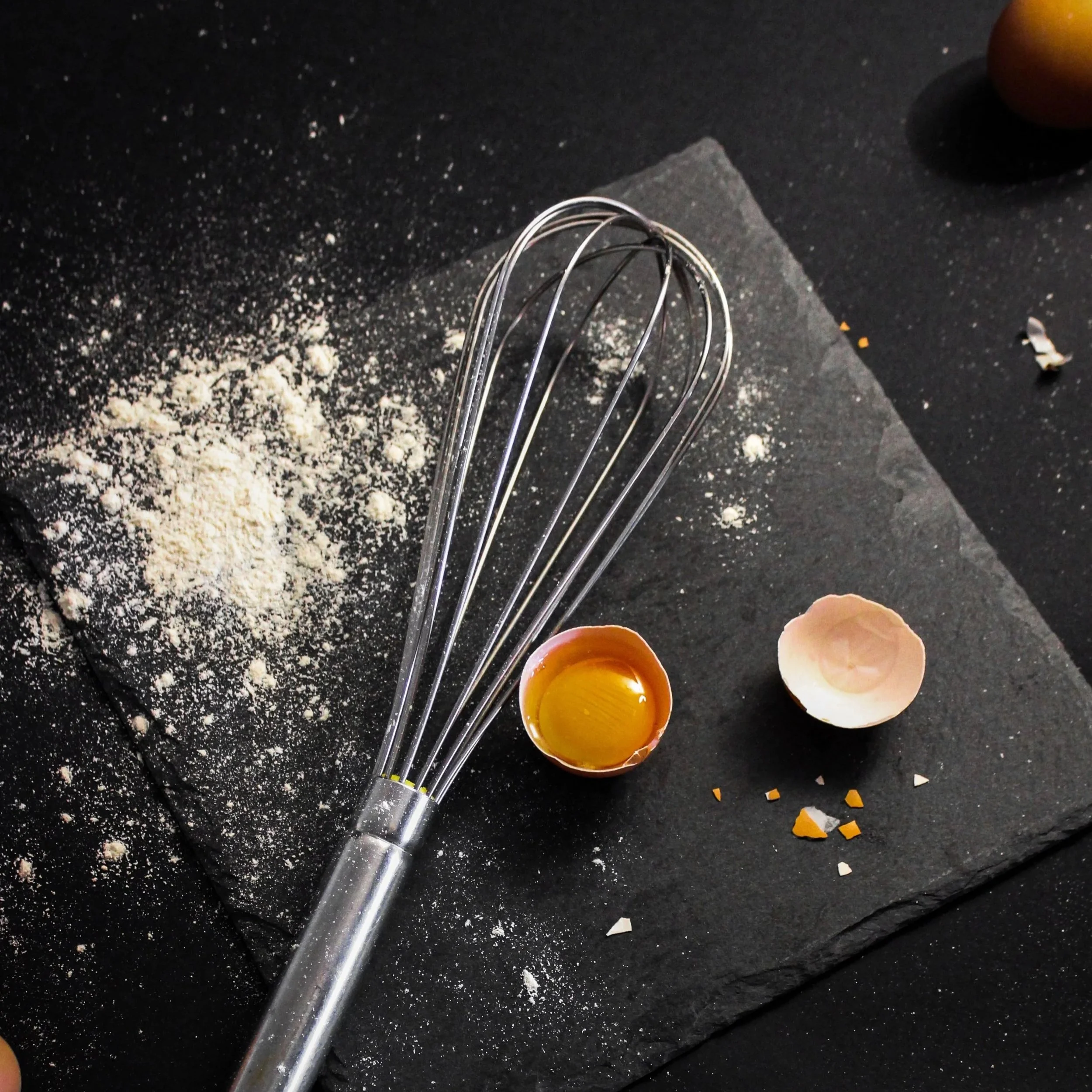 Baking scene with flour, eggs, eggshells, and a whisk on a dark countertop.