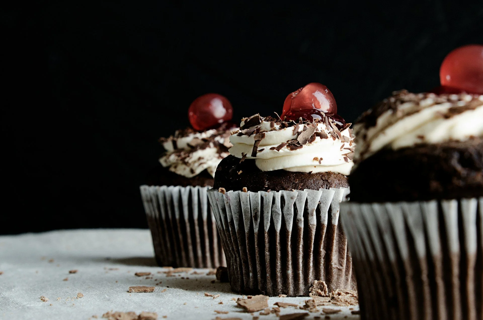 Three chocolate cupcakes topped with white buttercream, chocolate shavings, and cherries.