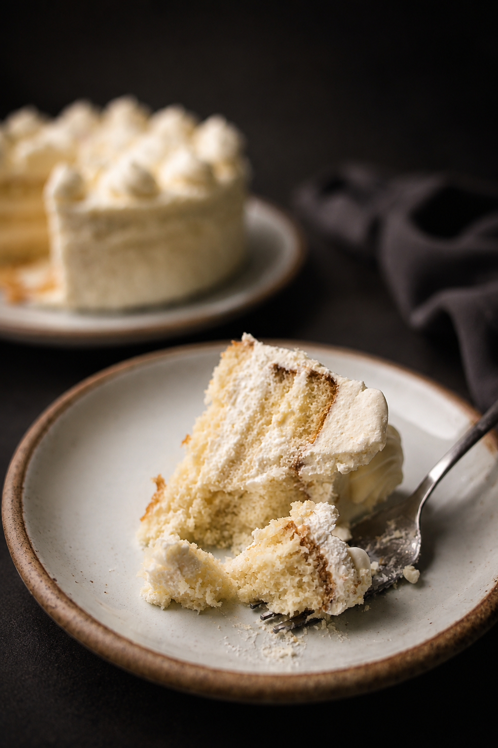 A slice of vanilla cake on a plate with a fork, with a full cake in the background.