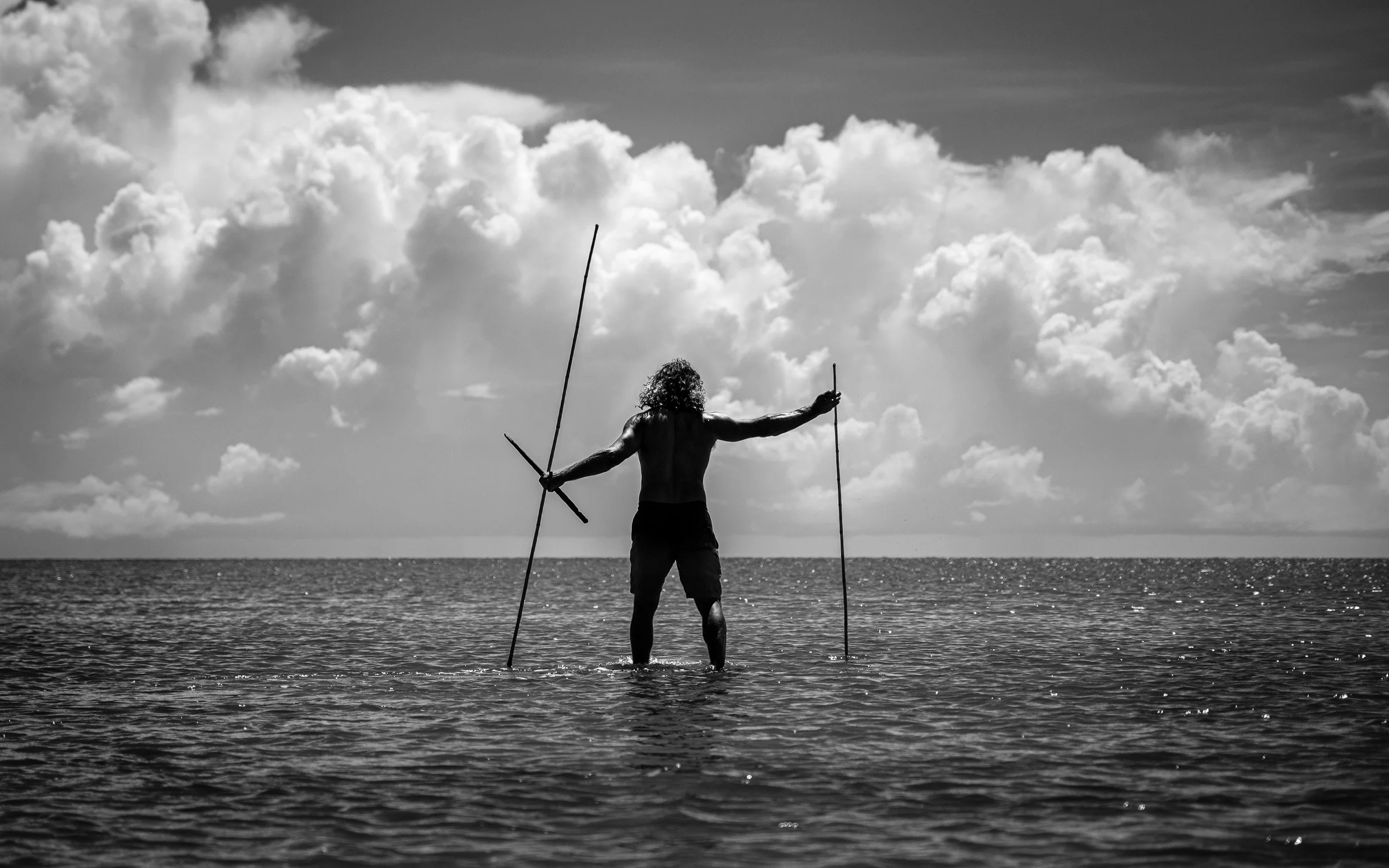 Black and white fine art photograph from the From Land to Sea series by Tommy Salpietro, depicting a Kuku Yalanji man standing in coastal waters with traditional fishing spears in Far North Queensland, Australia. Documentary photography exploring 