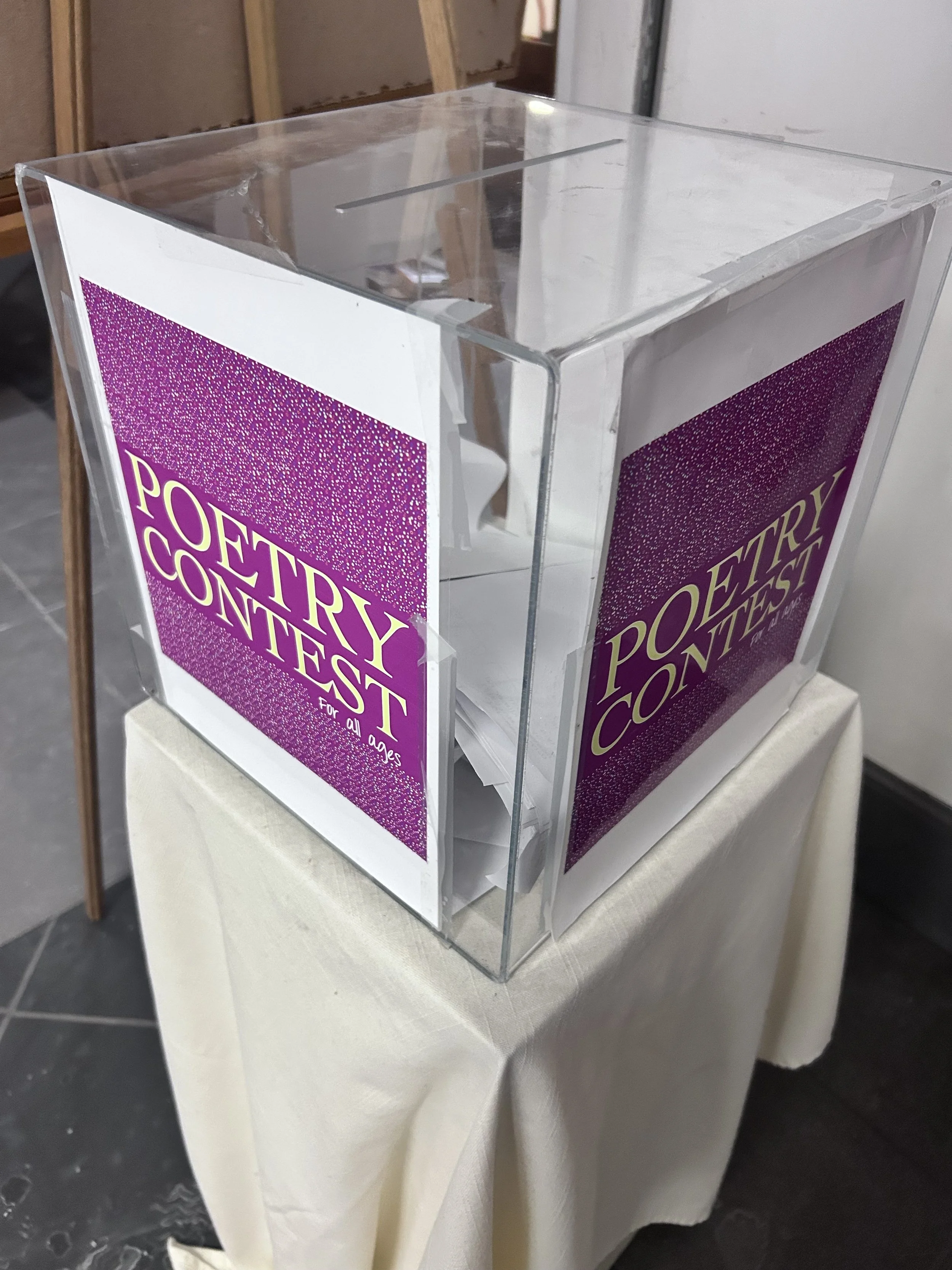 A transparent ballot box with purple and yellow 'Poetry Contest' posters on three sides. The box is placed on a white tablecloth-covered table, and the photo is taken indoors with a metal wall and unfinished wooden frame in the background.