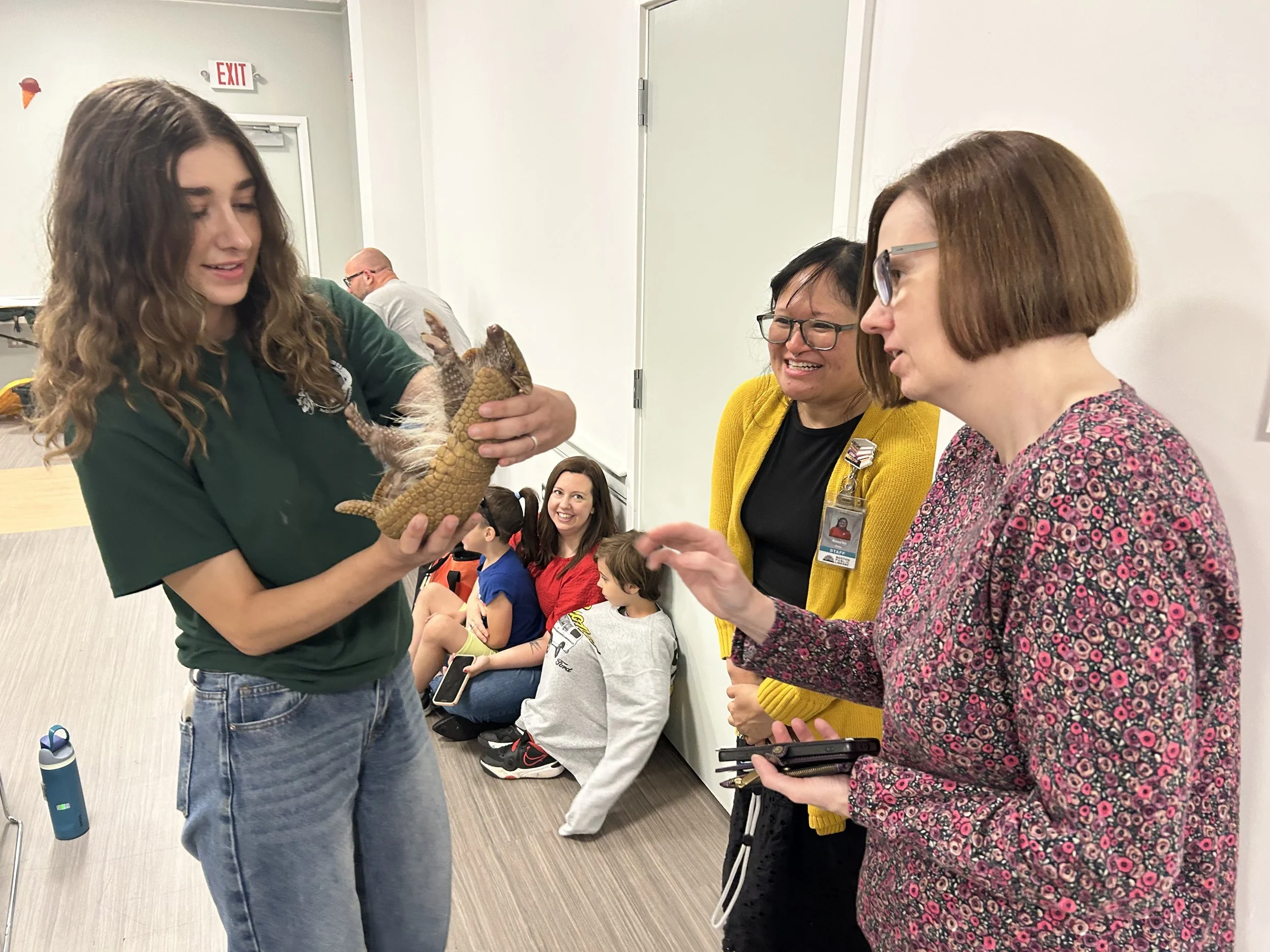 Three women are gathered in a room, engaging in an activity involving a small dinosaur plush toy. One woman with long curly hair is holding the plush dinosaur, while the two others with short hair and glasses look on and smile. Several children are seated against the wall in the background.