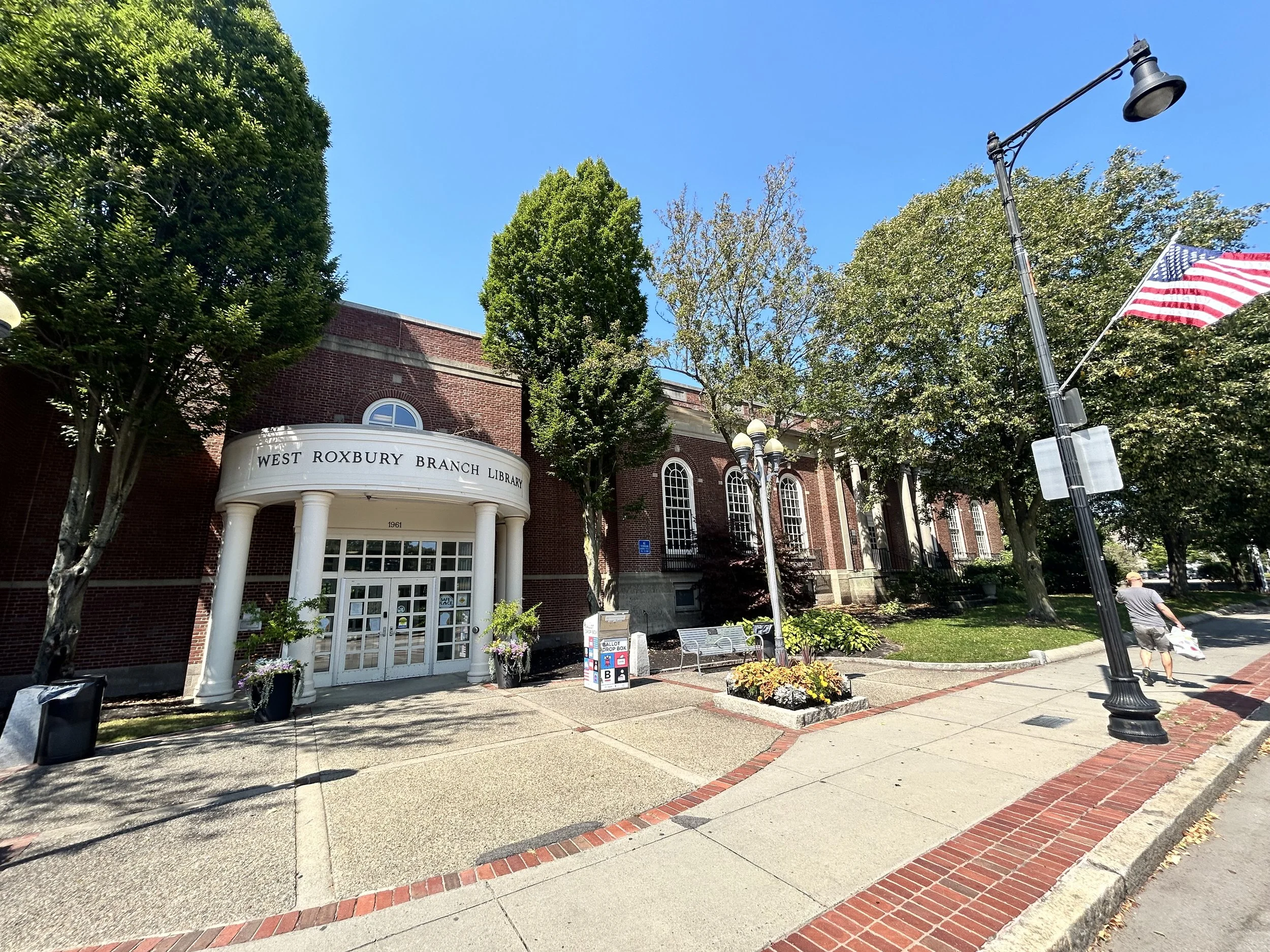 Exterior view of the West Roxbury Branch Library building with brick facade, white columns at the entrance, surrounded by trees, American flag, and streetlamp.