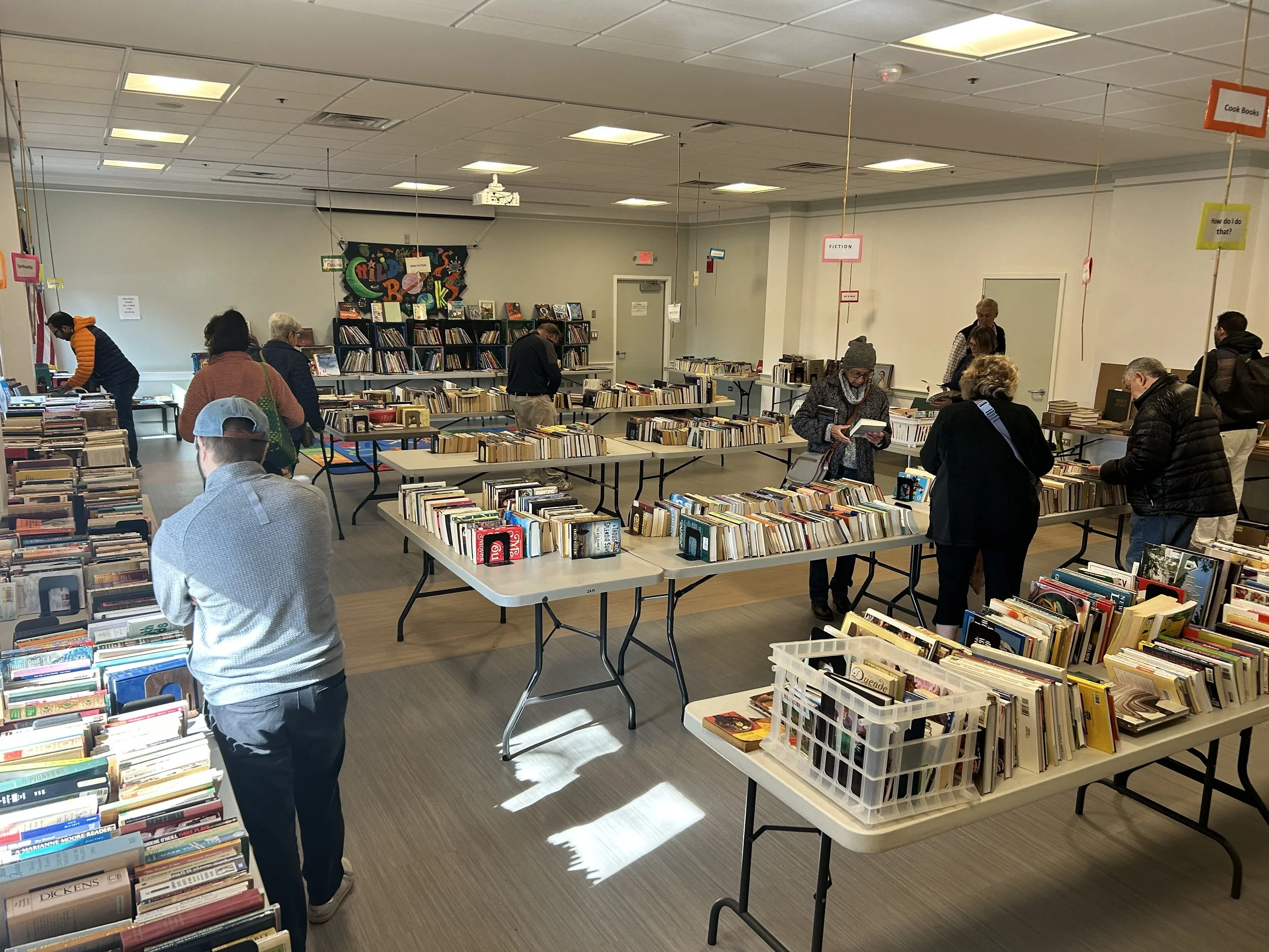 People browsing books at a book sale in a library or bookstore, with tables full of books and signage hanging from the ceiling.