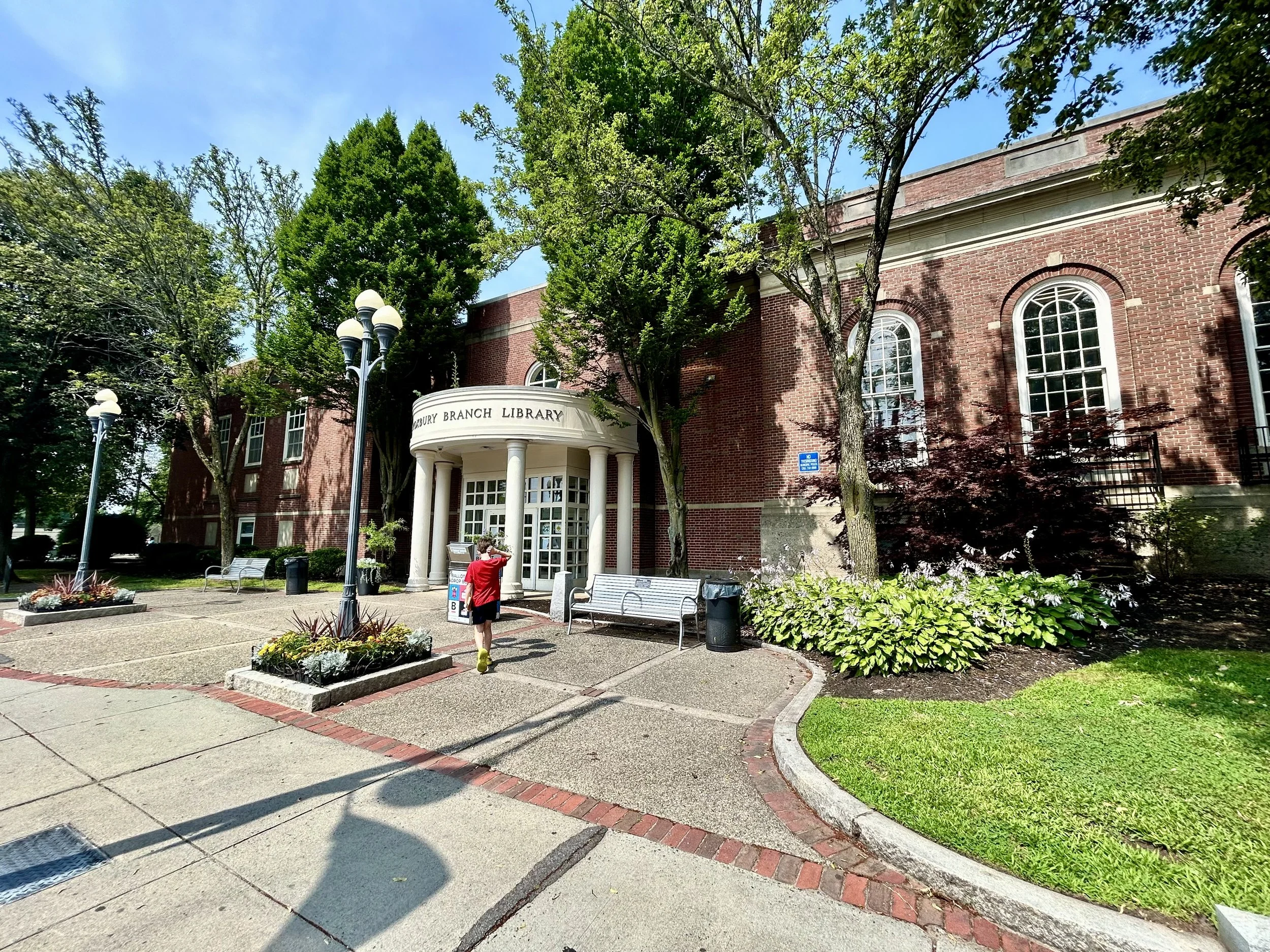 The exterior of a red brick library building with arched windows, surrounded by green trees, with a sign that reads 'Saxony Branch Library'. In front, a person in a red shirt is taking a photo, and there are benches and flower beds near the sidewalk.