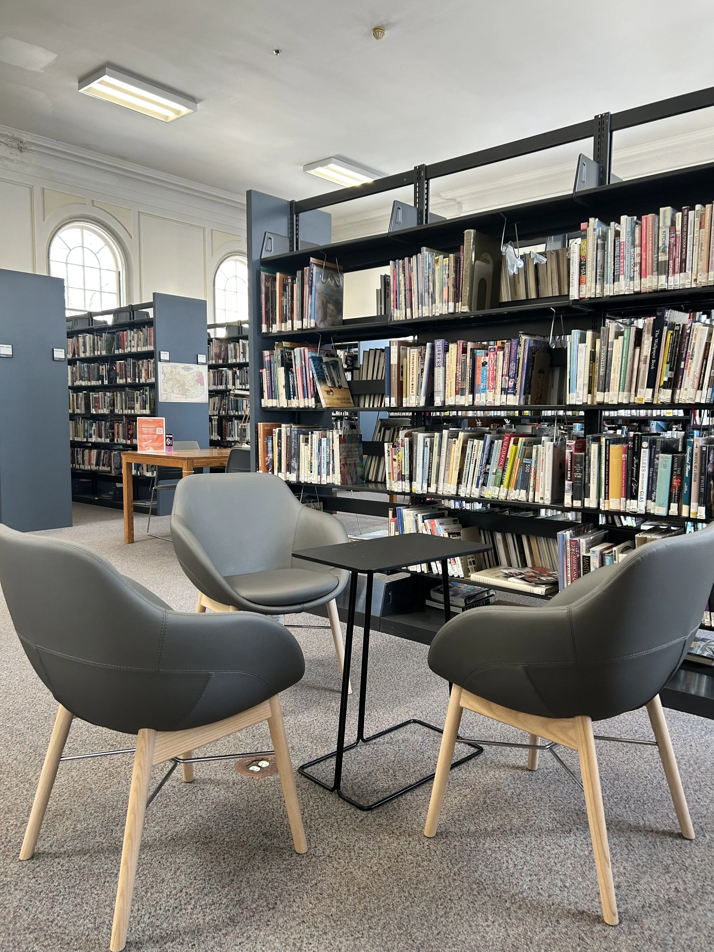 A cozy library area with four black chairs with wooden legs arranged around small tables, and bookshelves filled with books in the background.