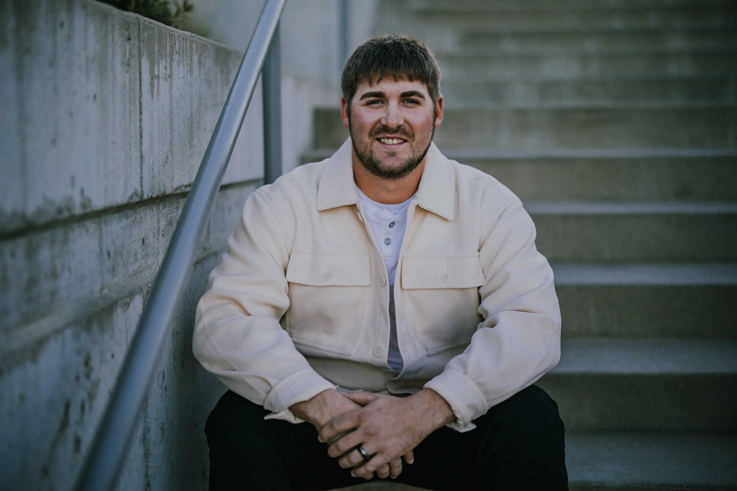 A man with dark hair and a beard sitting on concrete stairs, smiling, wearing a light-colored jacket and black pants