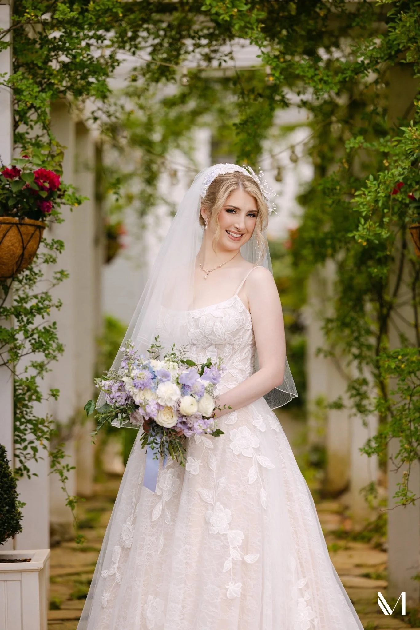 A smiling bride with blonde hair in an elegant white lace wedding gown, holding a bouquet of white, purple, and blue flowers, standing outdoors under an archway decorated with greenery and hanging plants.