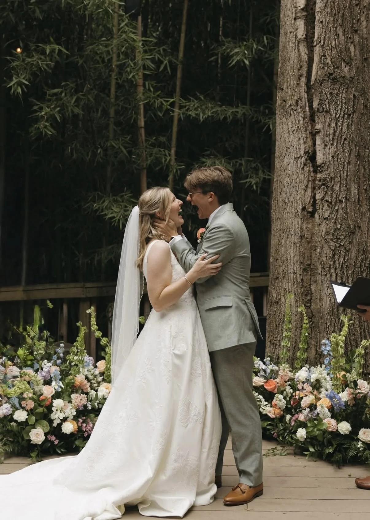 A bride and groom exchanging vows during a wedding ceremony outdoors. The bride is in a white wedding dress with a veil, and the groom is in a light gray suit. They are smiling and facing each other close, surrounded by flowers and greenery.