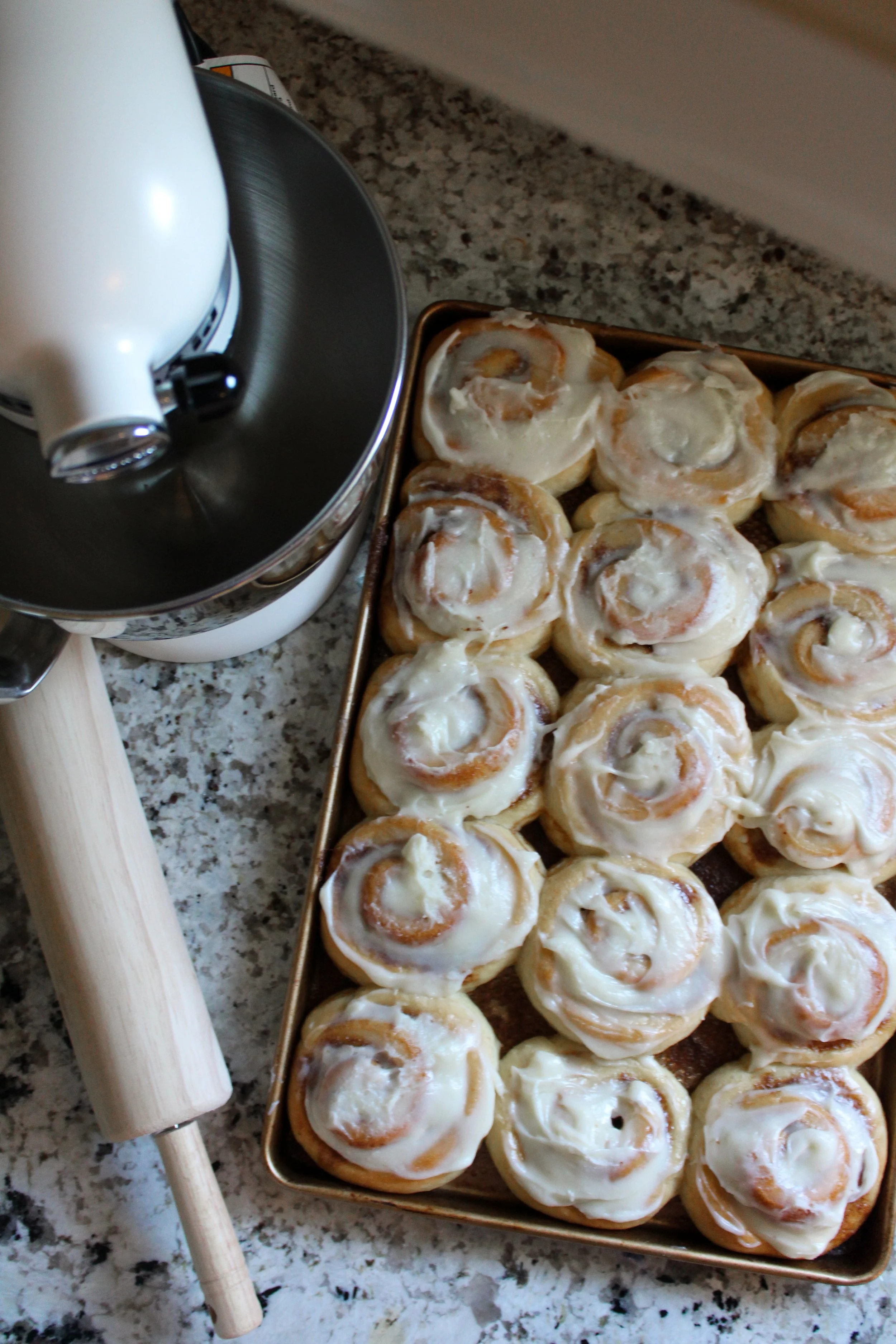 A baking sheet filled with cinnamon rolls topped with icing, a rolling pin, and an electric hand mixer on a granite countertop.