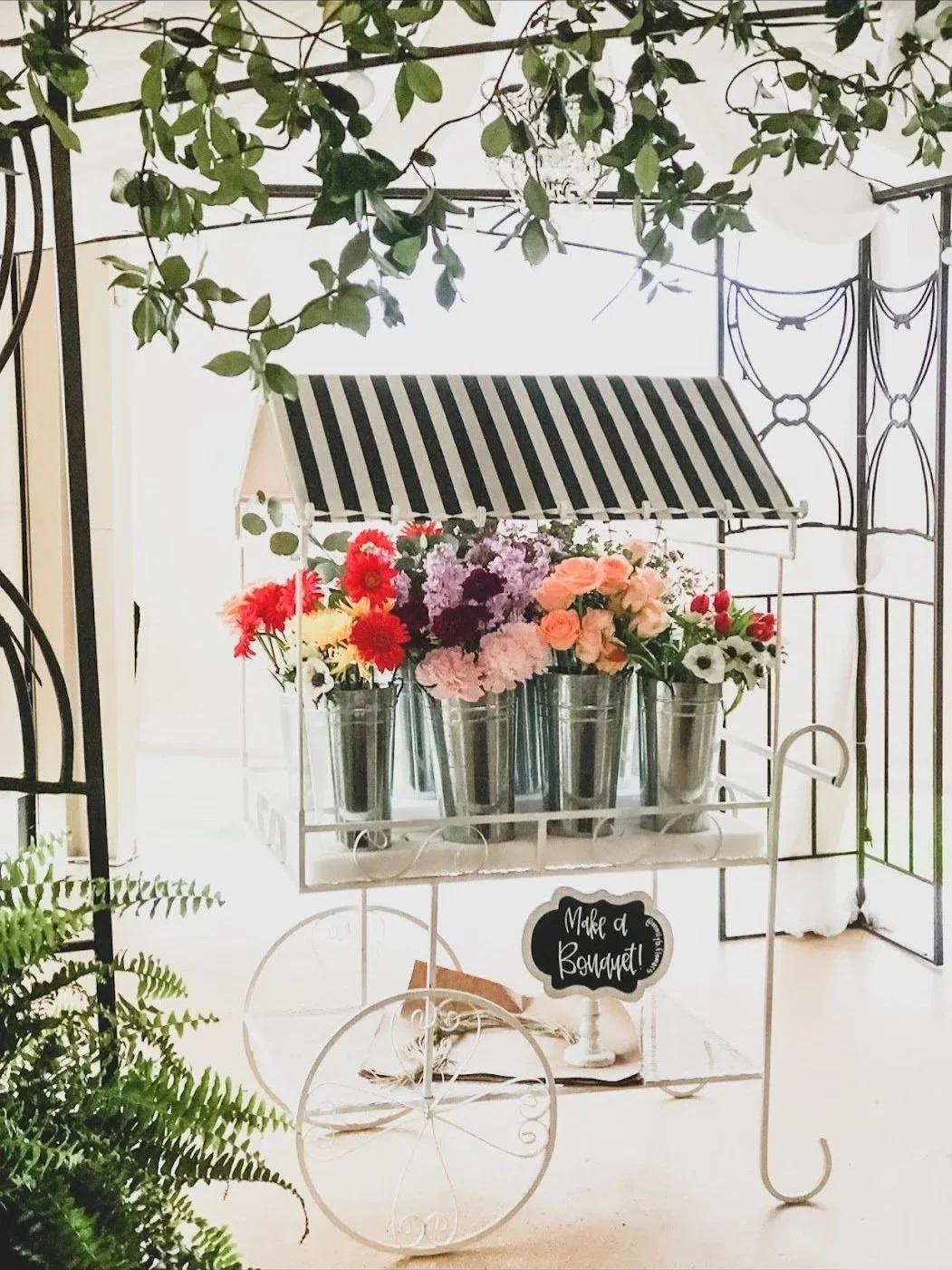 A decorative white metal cart with flowers in metal vases, a striped canopy, and a chalkboard sign that reads "Make a Bouquet!" set in a bright, airy space with green plants and black wrought iron decor.