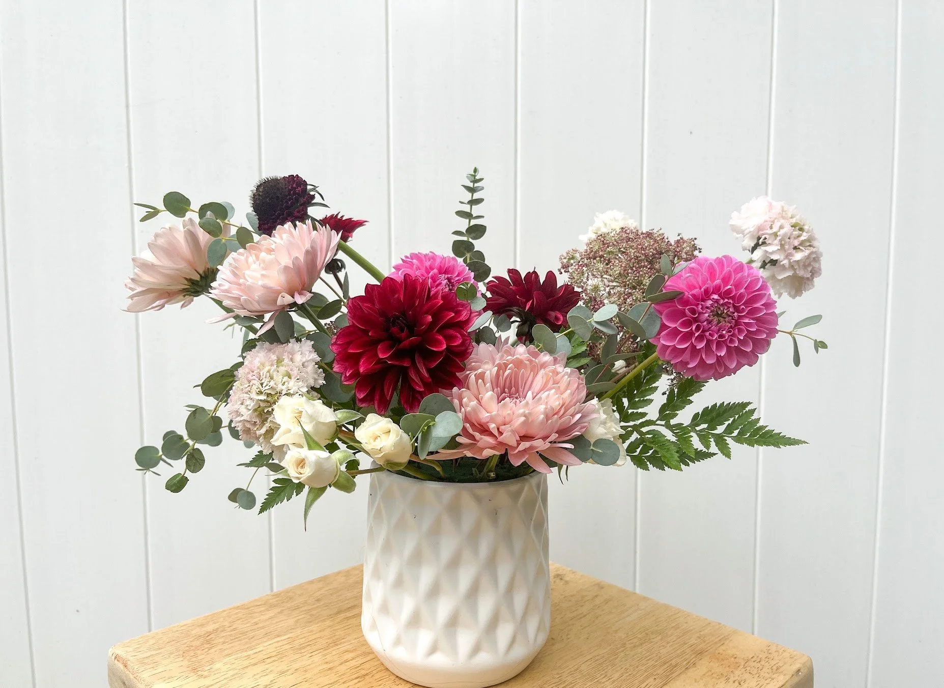 A bouquet of pink, white, and dark red flowers, including dahlias and roses, arranged with green foliage and placed in a white textured ceramic vase on a wooden surface, with a white paneled wall in the background.