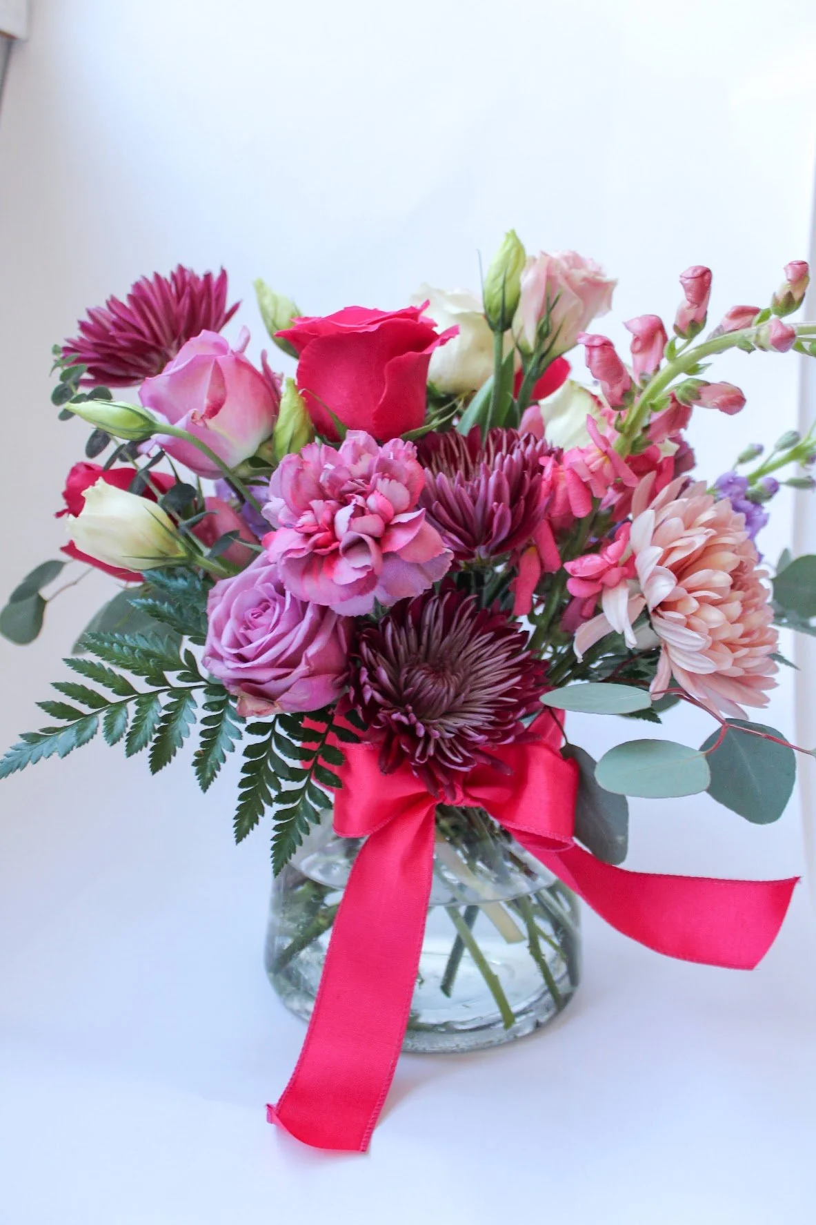 A bouquet of pink, red, and white flowers in a glass vase with a pink ribbon tied around it, set against a white background.