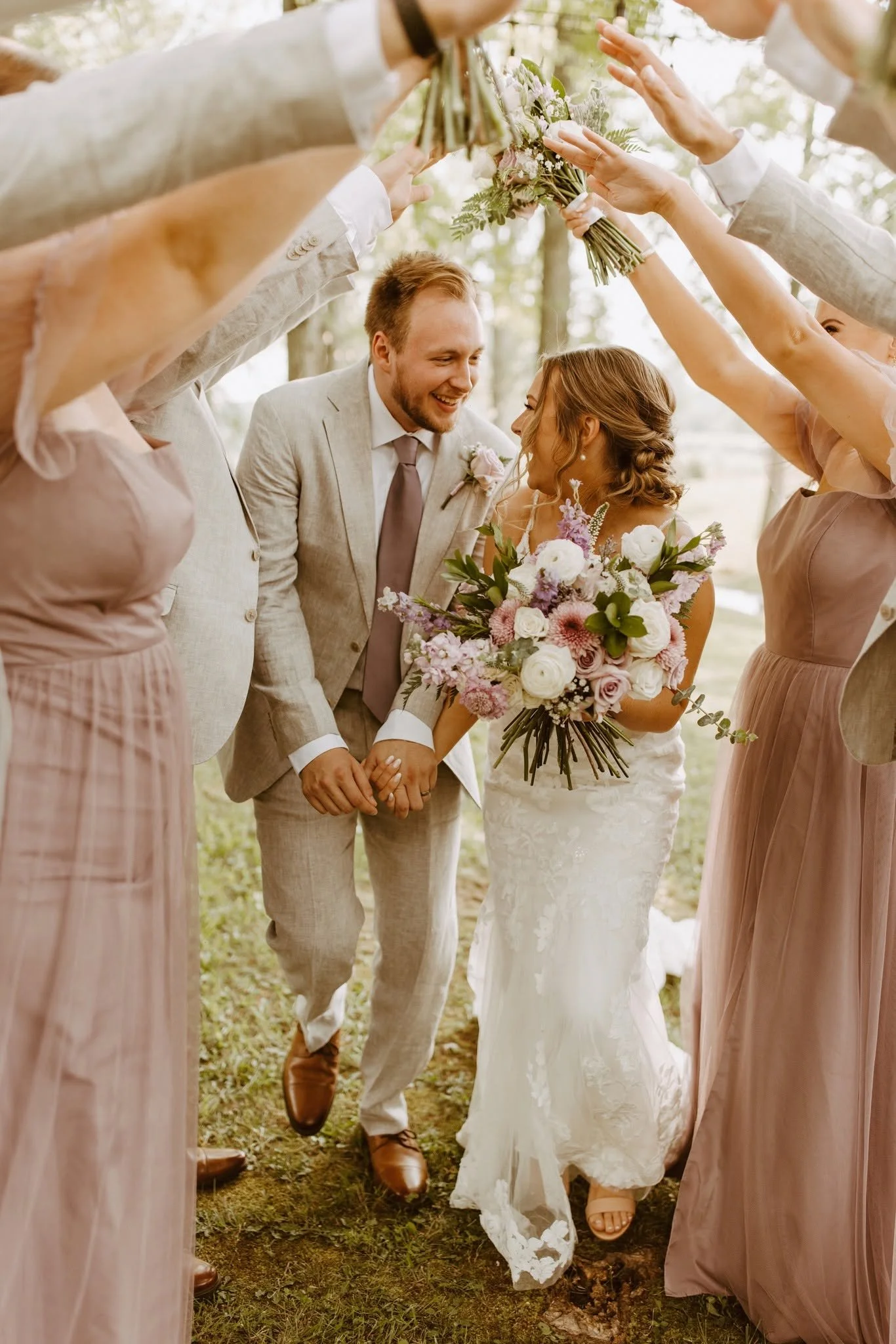 A newlywed couple holding hands and smiling under a canopy of friends at an outdoor wedding ceremony, surrounded by bridesmaids and friends, with the bride holding a large bouquet of flowers.