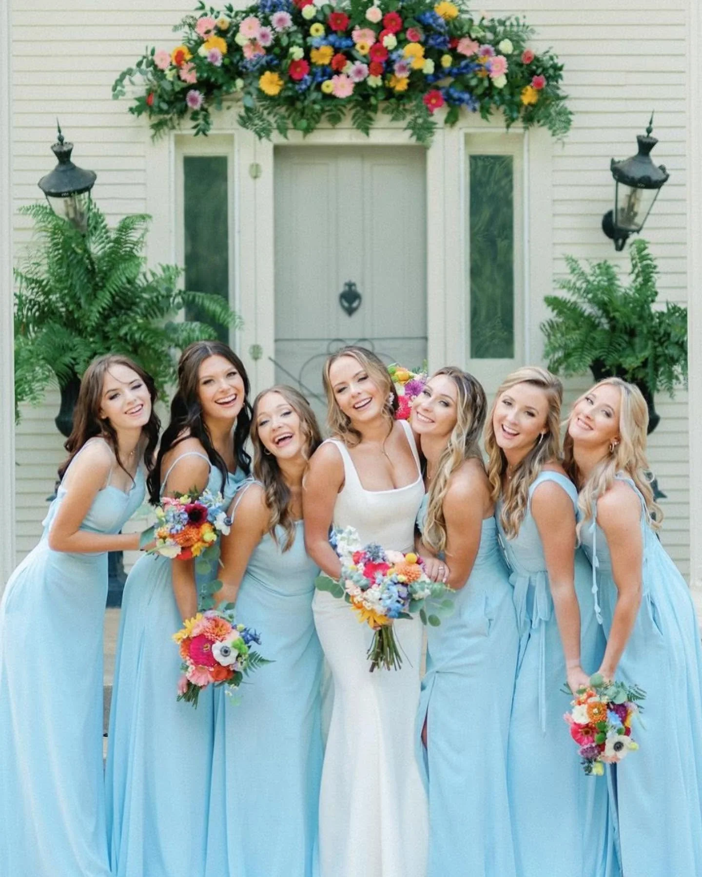 Group of bridesmaids in light blue dresses holding colorful bouquets, standing in front of a house with floral decorations.