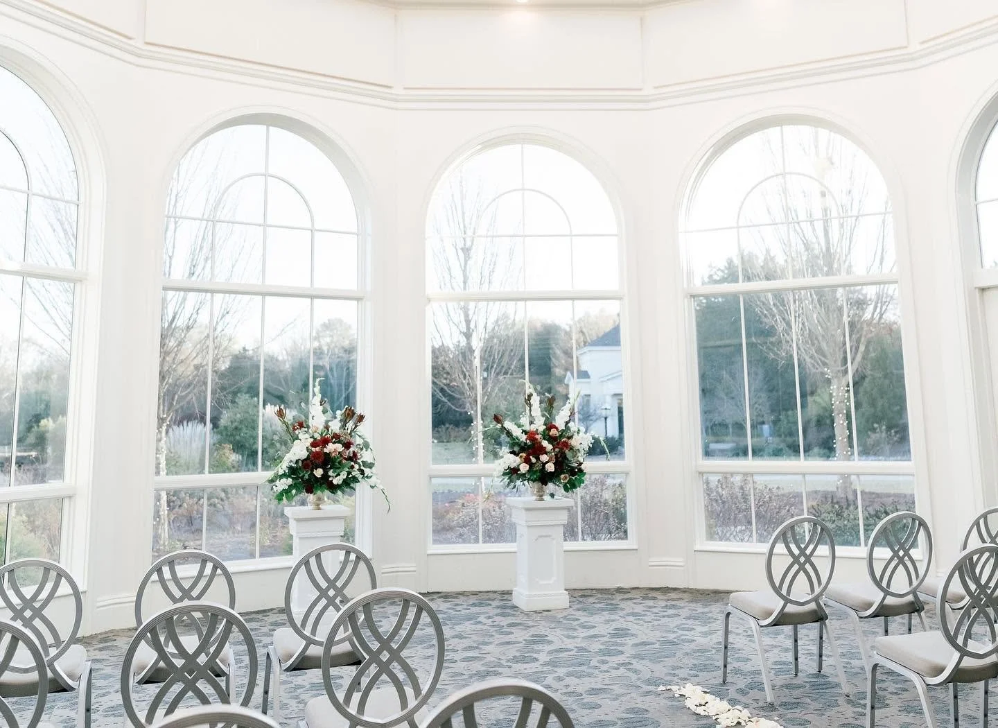 A bright interior room with large arched windows, white walls, floral arrangements on white pedestals, and empty chairs arranged in a semi-circle, suggesting a setup for a wedding or special event.