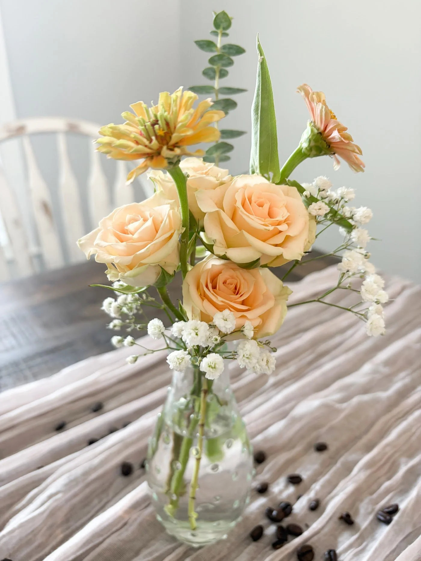 A glass vase with a floral arrangement of peach roses, pink and yellow zinnias, white baby's breath, and greenery, placed on a wooden surface with scattered coffee beans, against a light background.
