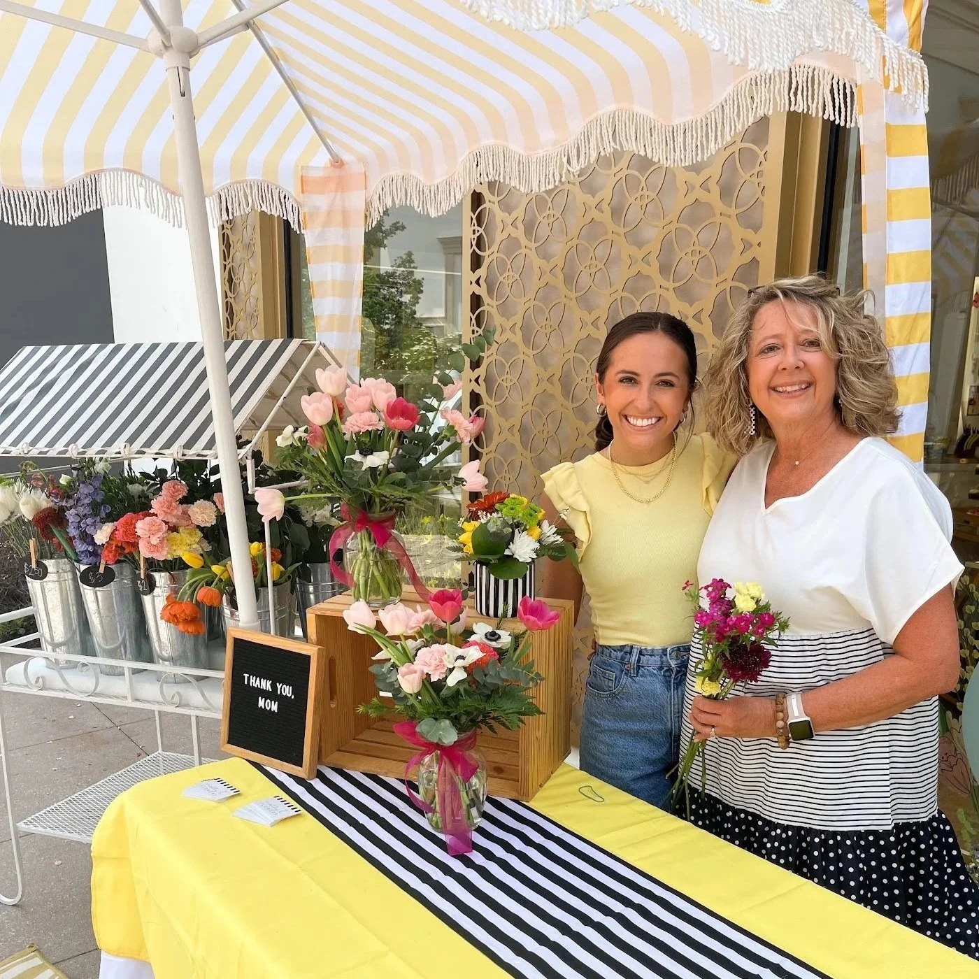 Two women standing behind a flower display at a stall with yellow and black striped tablecloth and black and white striped decorations. One woman holds a small bouquet of flowers; the other stands next to her. There is a sign on the table that reads "Thank you, mom." and vases with pink, white, purple, yellow, and orange flowers.