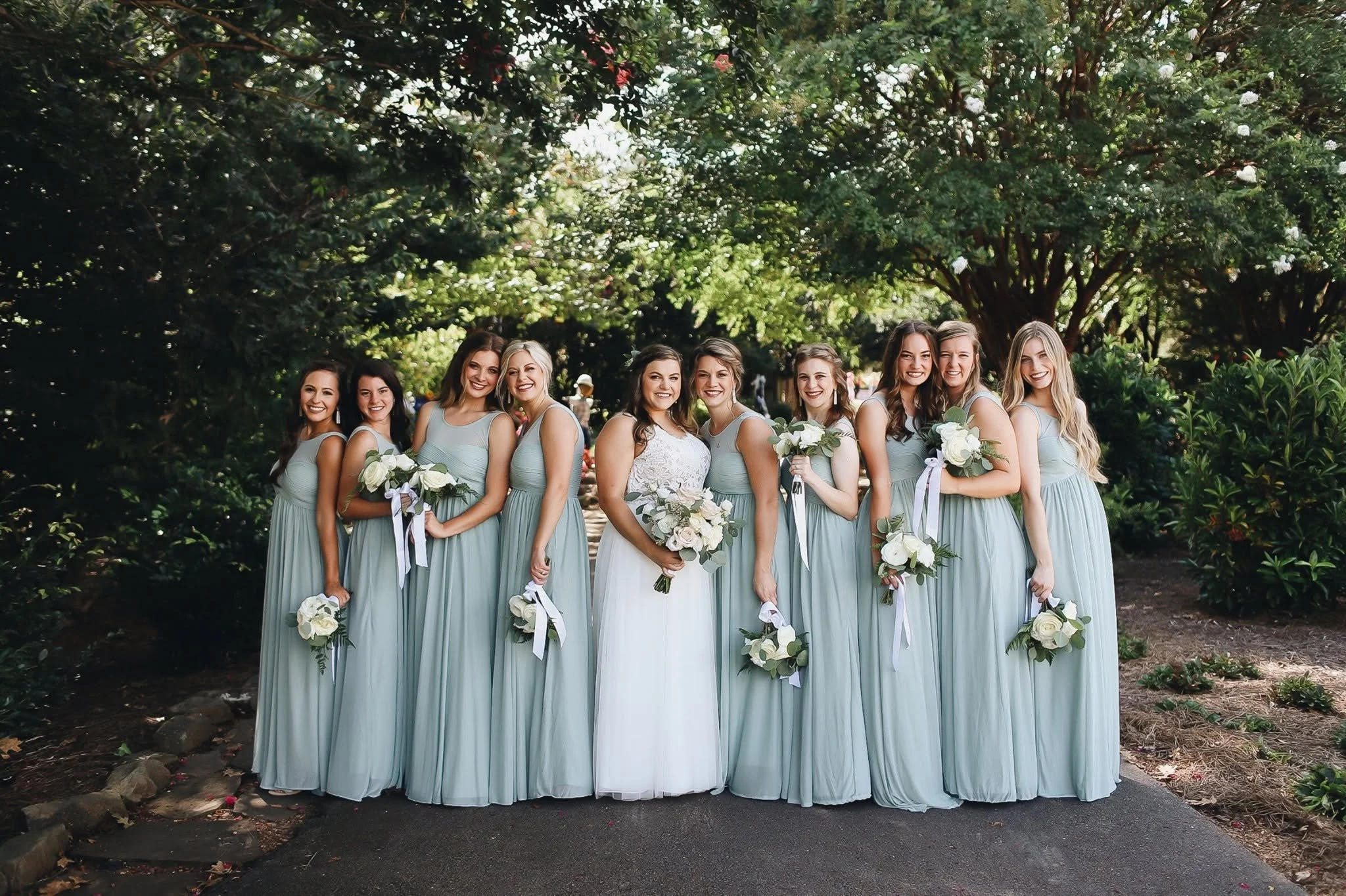 Group of women in matching light blue bridesmaid dresses standing outdoors under trees, smiling, some holding bouquets of white flowers.