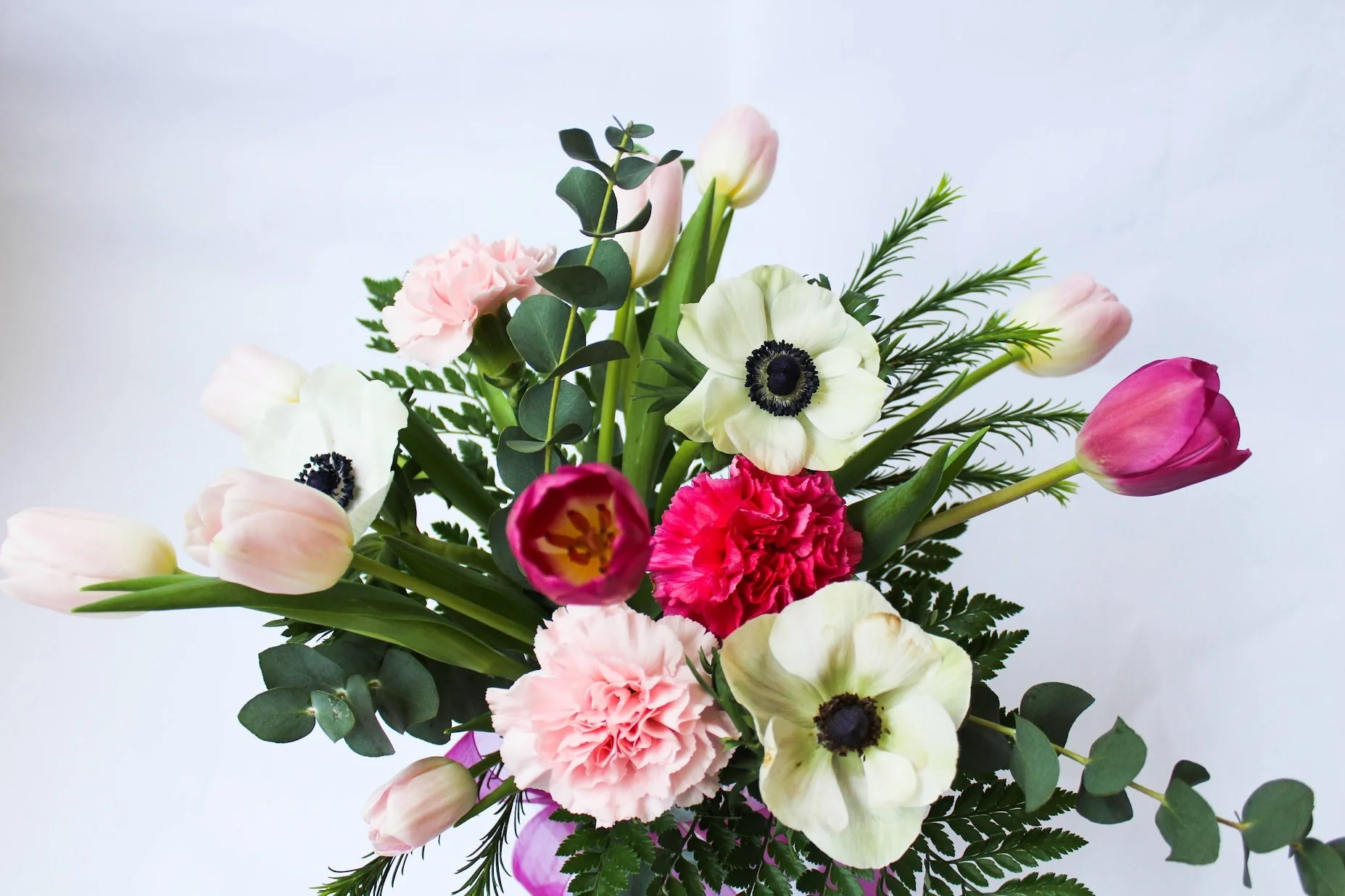 A bouquet of various flowers including pink tulips, white poppies, pink carnations, and greenery against a white background.