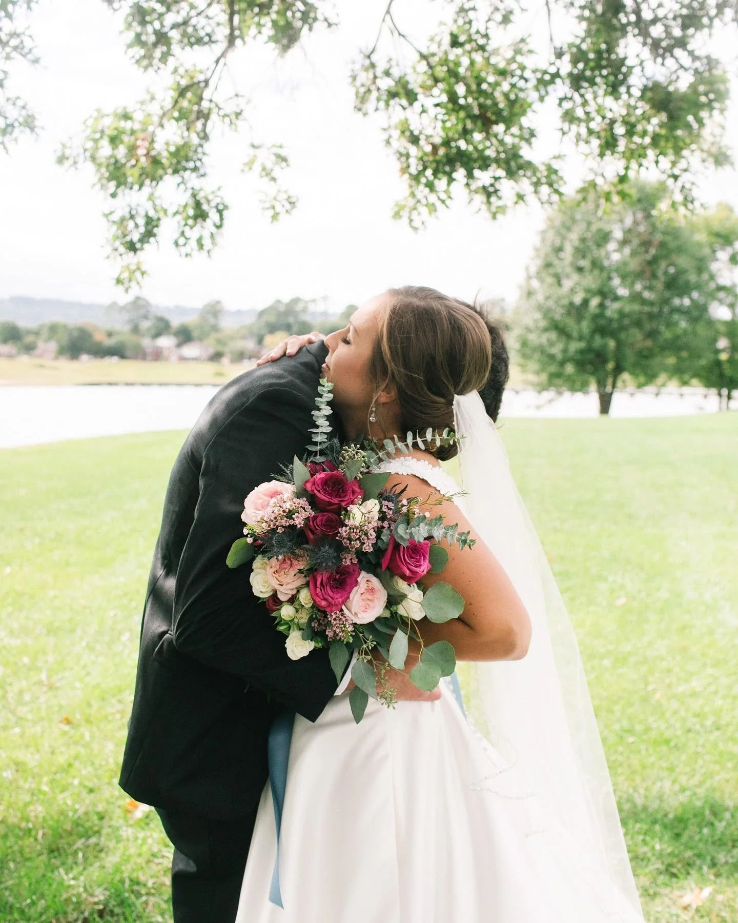 A bride and groom embrace outdoors near a lake with trees in the background, the bride holding a large bouquet of pink, white, and purple flowers.