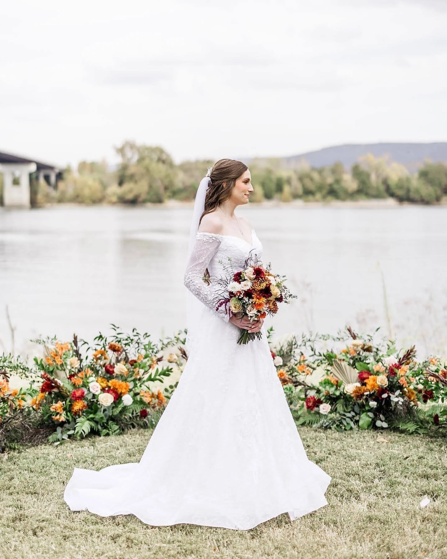 Bride in a white lace wedding dress holding a bouquet of flowers, standing outdoors near a river with trees and mountains in the background, surrounded by a floral arrangement.