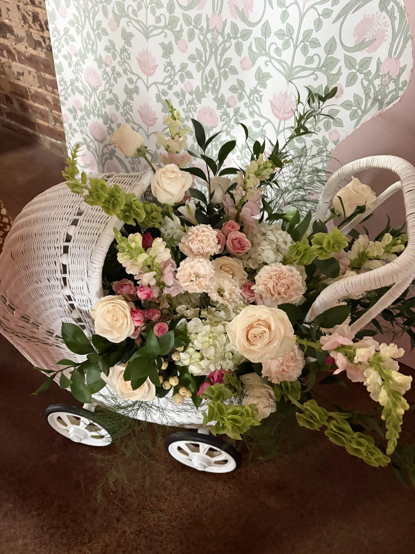 A white wicker cart filled with a variety of pink, white, and cream flowers, including roses and carnations, with green foliage, set against a wallpapered wall with pink floral patterns and a brick wall in the background.