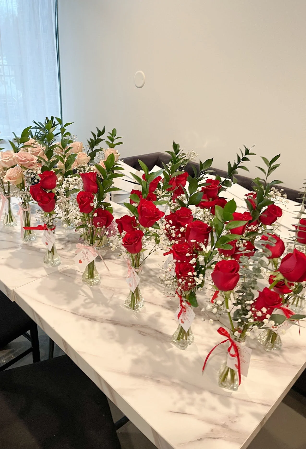 Multiple bouquets of red and pink roses with white baby's breath in glass vases on a white marble table against a plain wall.