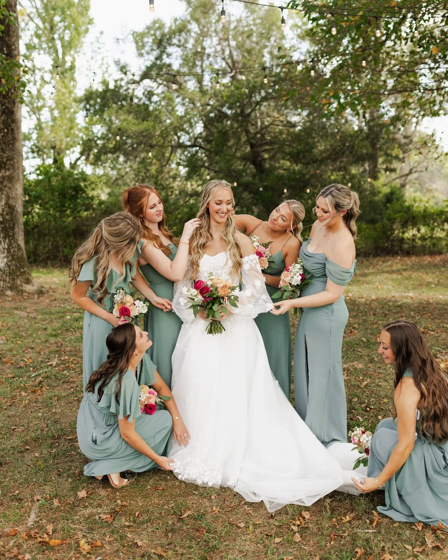 A bride in a white wedding gown surrounded by six bridesmaids in light teal dresses, outdoors under string lights, with trees in the background, as they prepare for a wedding photo.