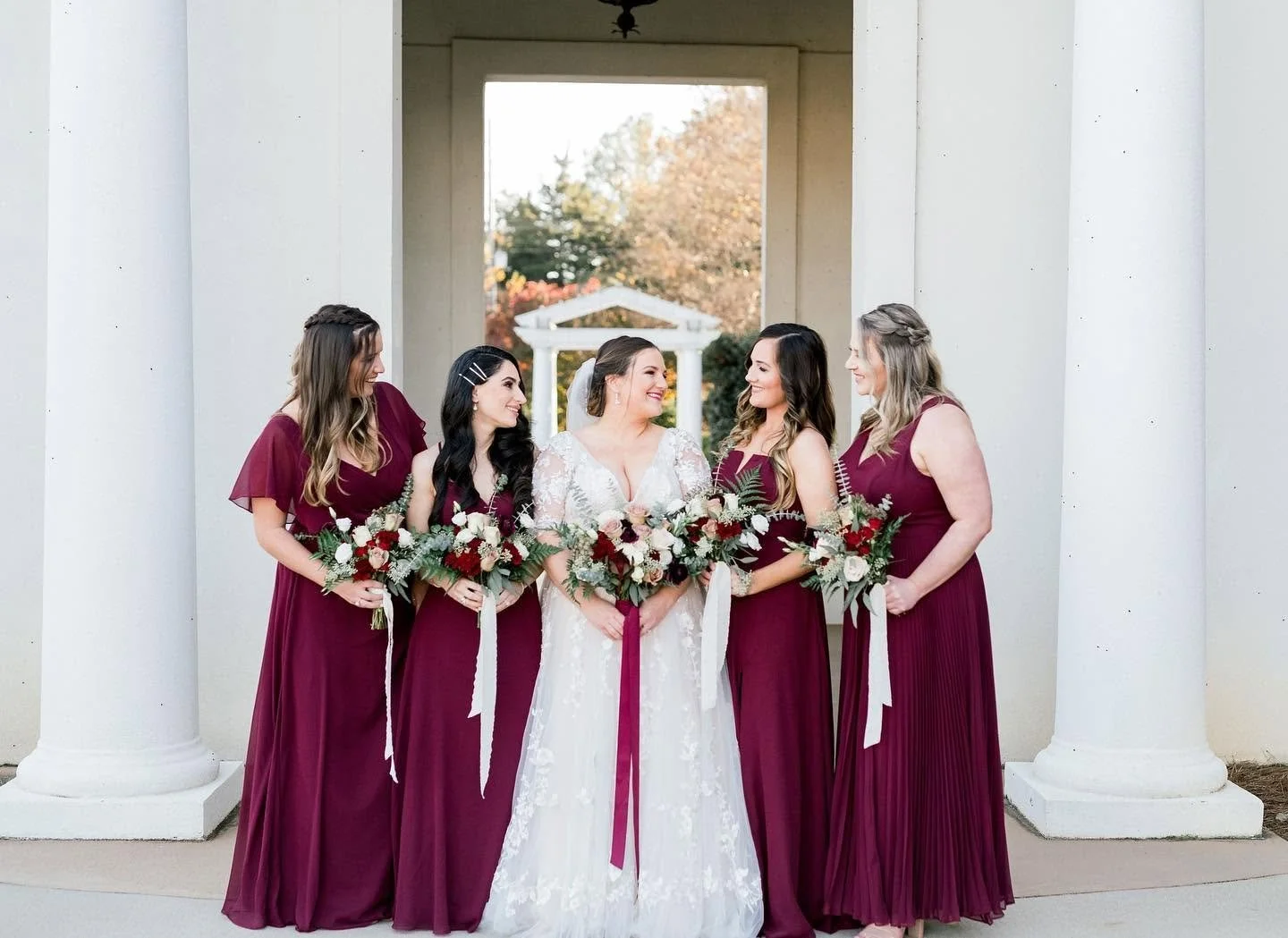 Bride and four bridesmaids in deep red dresses holding bouquets, standing under white columns outdoors.